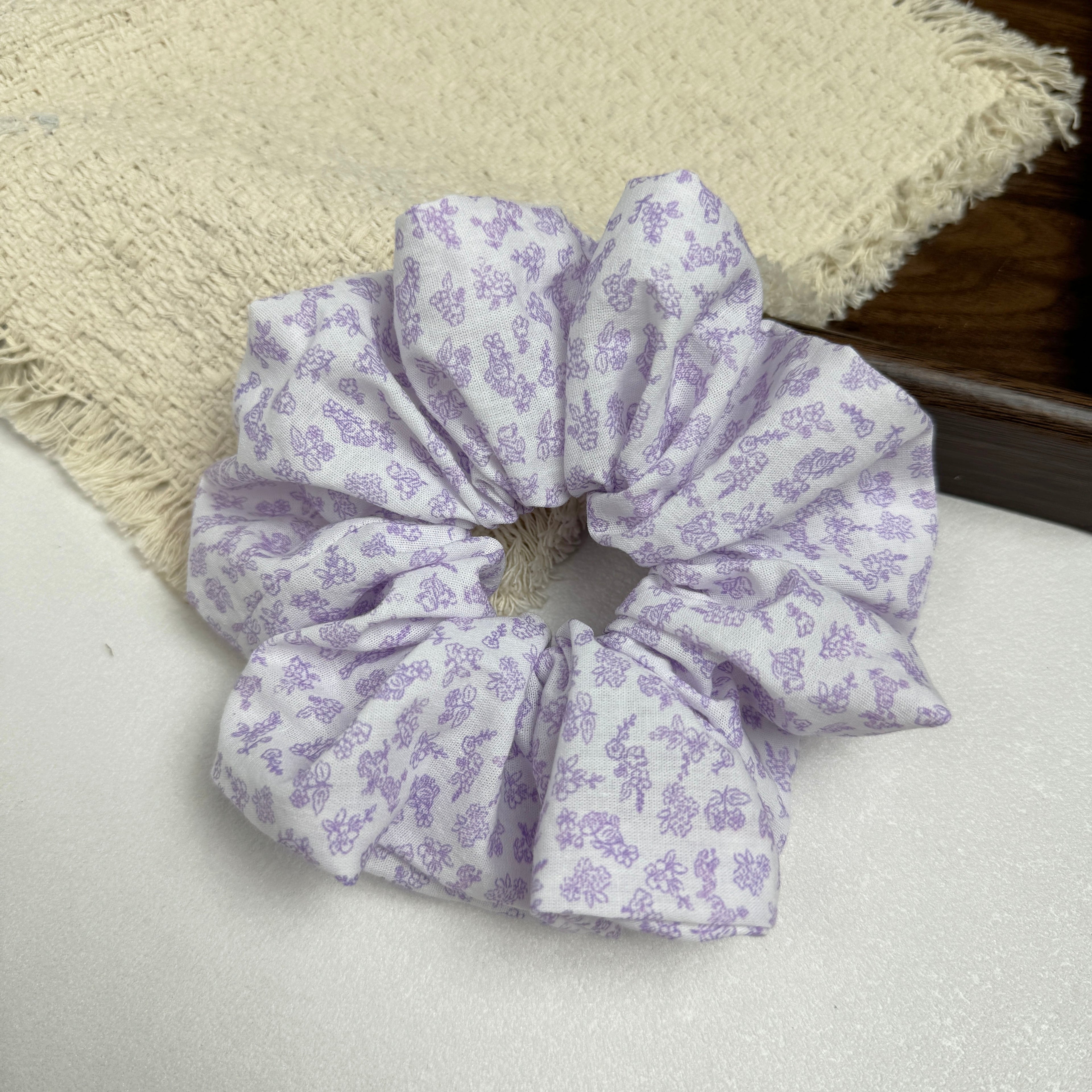 White lavender floral print scrunchie on a white surface beside a frayed cream woven fabric and the corner of a dark brown wooden tray, flat-lay composition.
