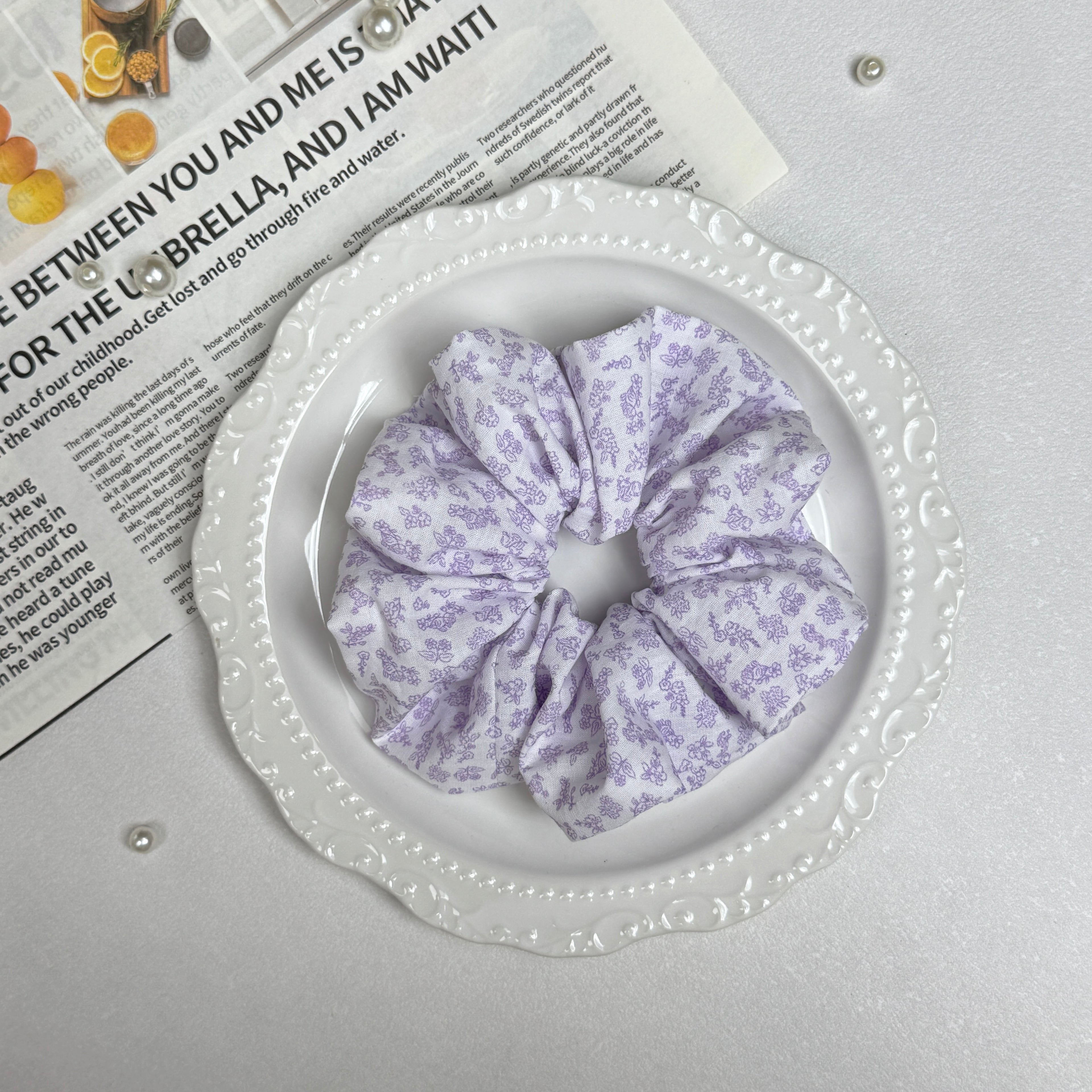 White lavender floral print scrunchie placed on a white ceramic plate with embossed scalloped rim, resting on a magazine page with citrus photography and text, scattered white faux pearls, grey-white surface.
