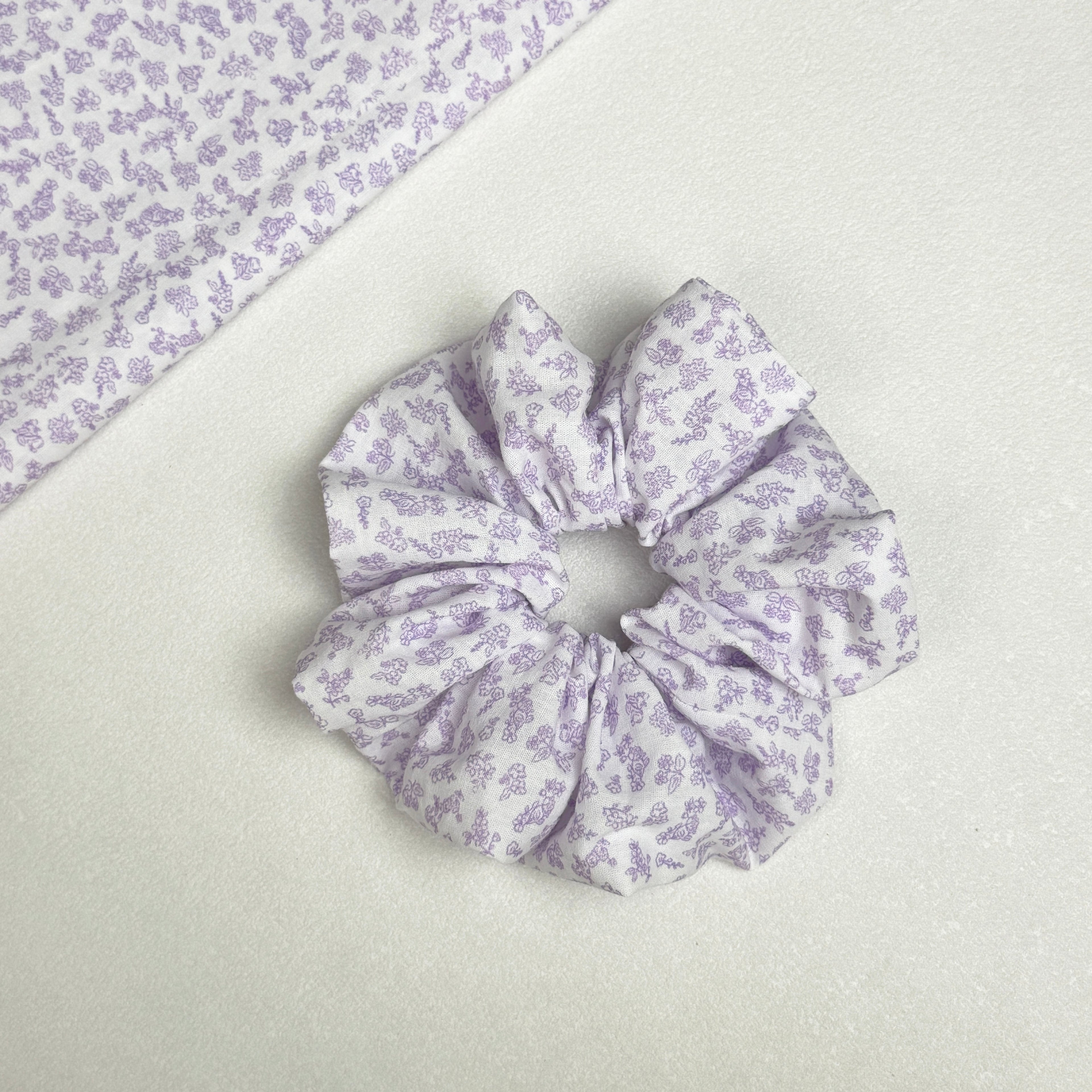 White lavender floral print scrunchie displayed alongside a matching fabric swatch laid diagonally, showing the full fine-line floral pattern, white pebbled texture surface.