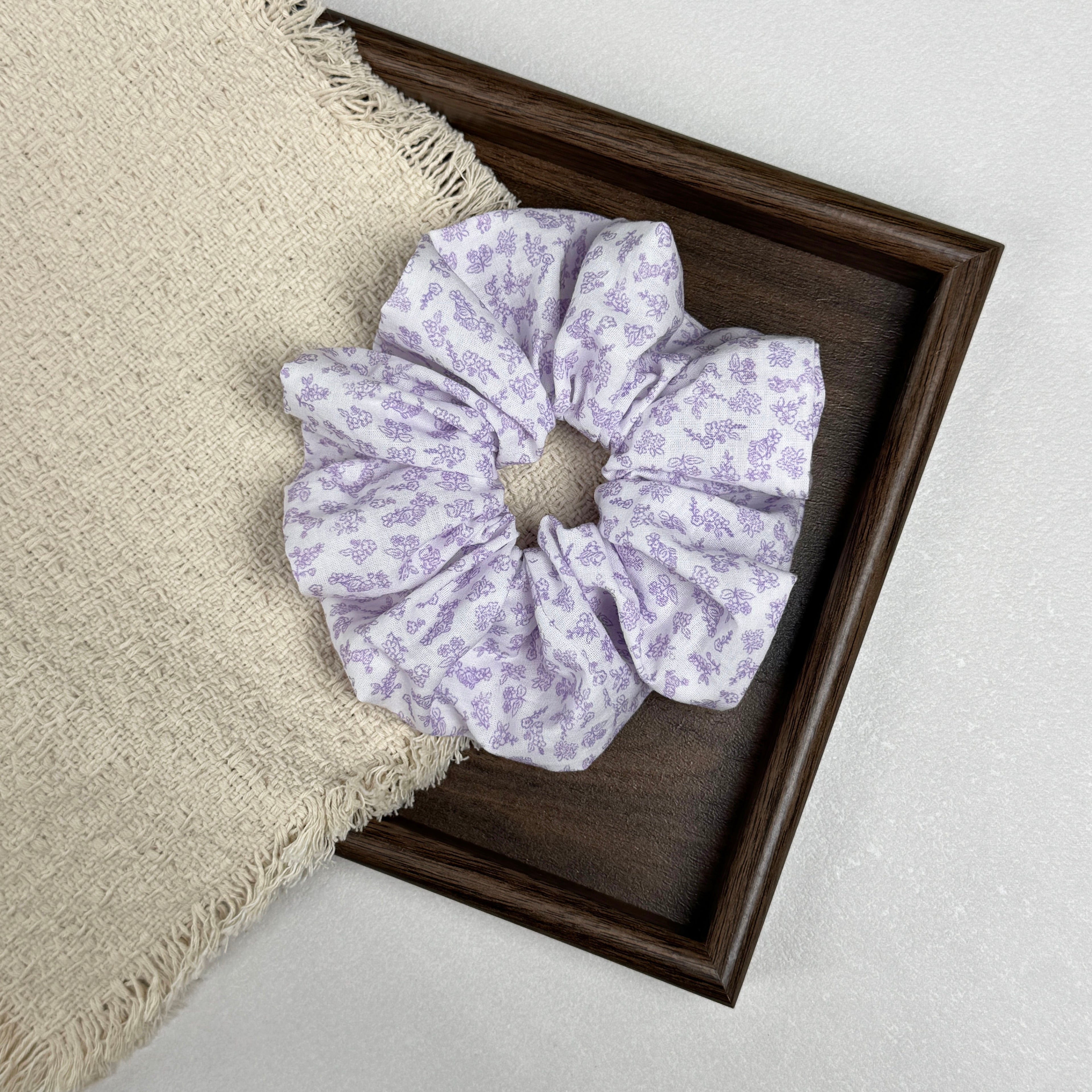 White lavender floral print scrunchie flat-laid across a dark brown wooden tray and a fringed cream woven fabric with shaggy tasseled edges, grey-white textured surface.