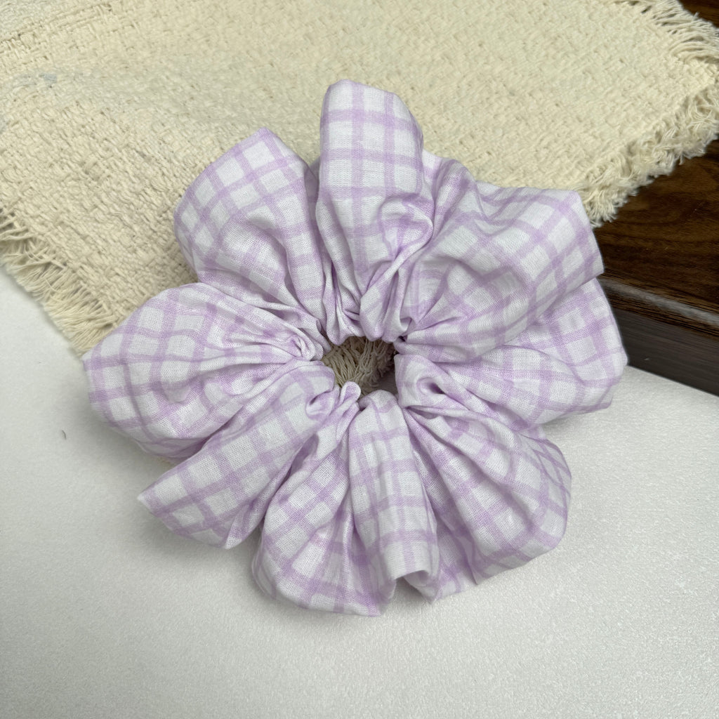 Lavender windowpane plaid scrunchie on a white surface with a frayed cream woven fabric on the left and dark brown wood on the right, flat-lay composition.