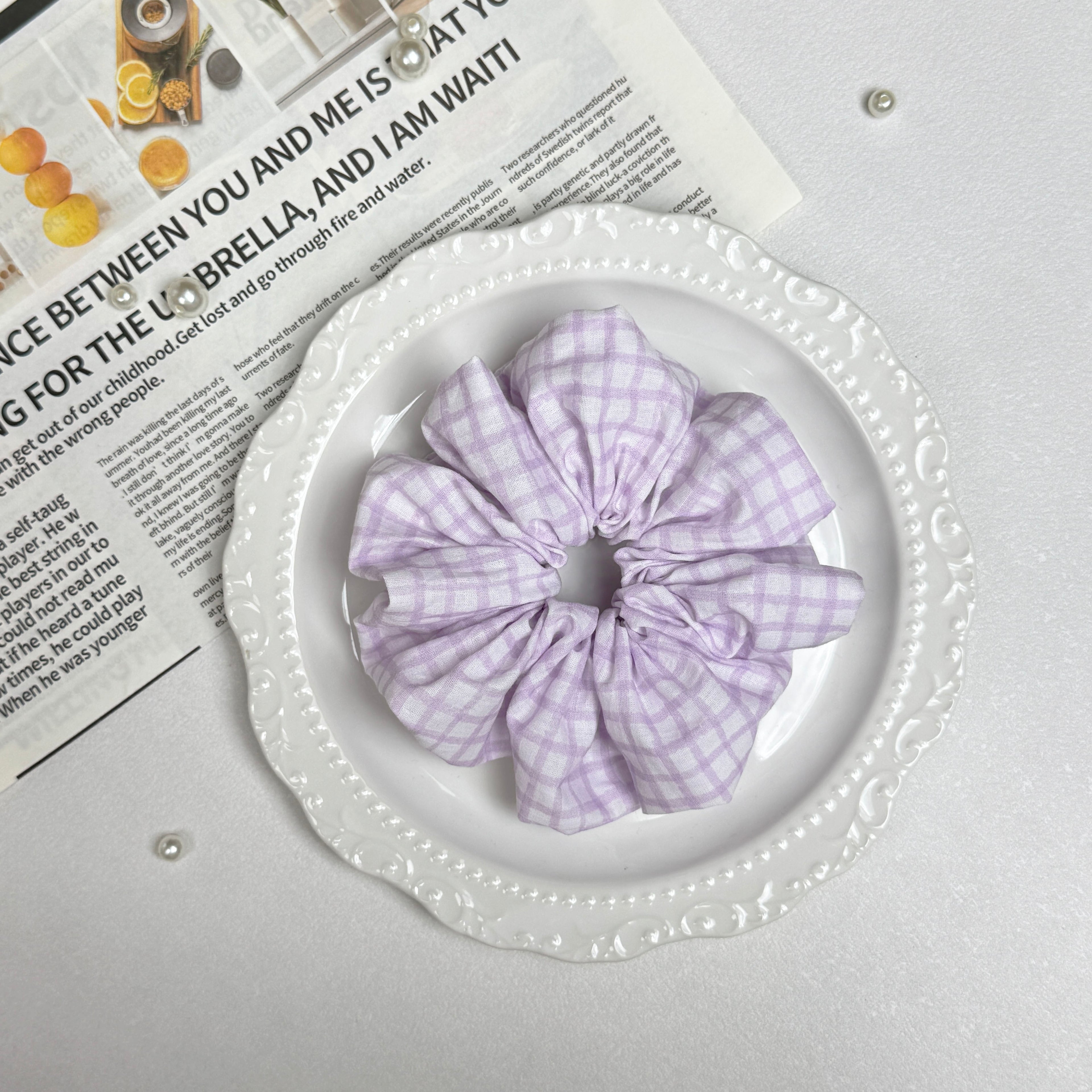 Lavender windowpane plaid scrunchie placed on a white ceramic plate with embossed scalloped rim, resting on a magazine page with citrus photography and text, scattered white faux pearls, grey-white surface.