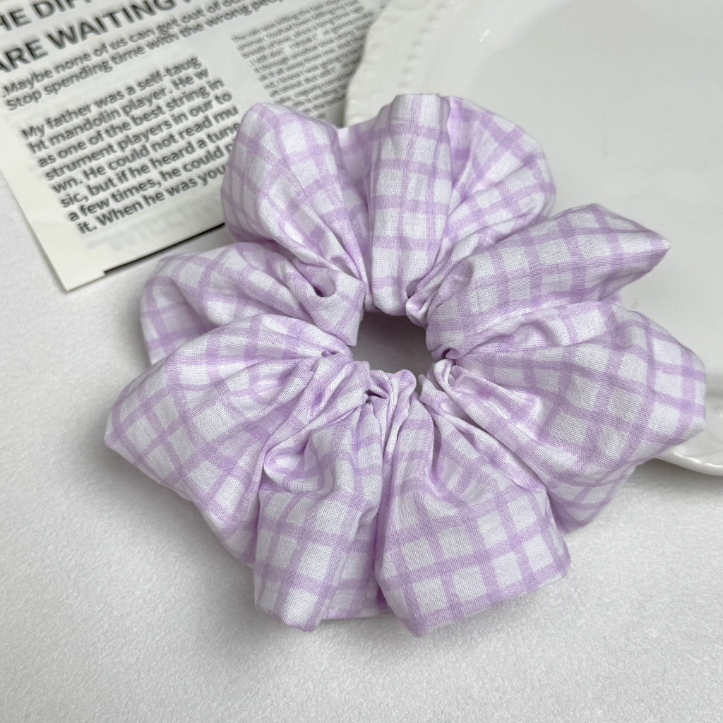 Lavender windowpane plaid scrunchie alongside a white ceramic plate with beaded scalloped rim and an open magazine page with printed text, flat-lay composition.