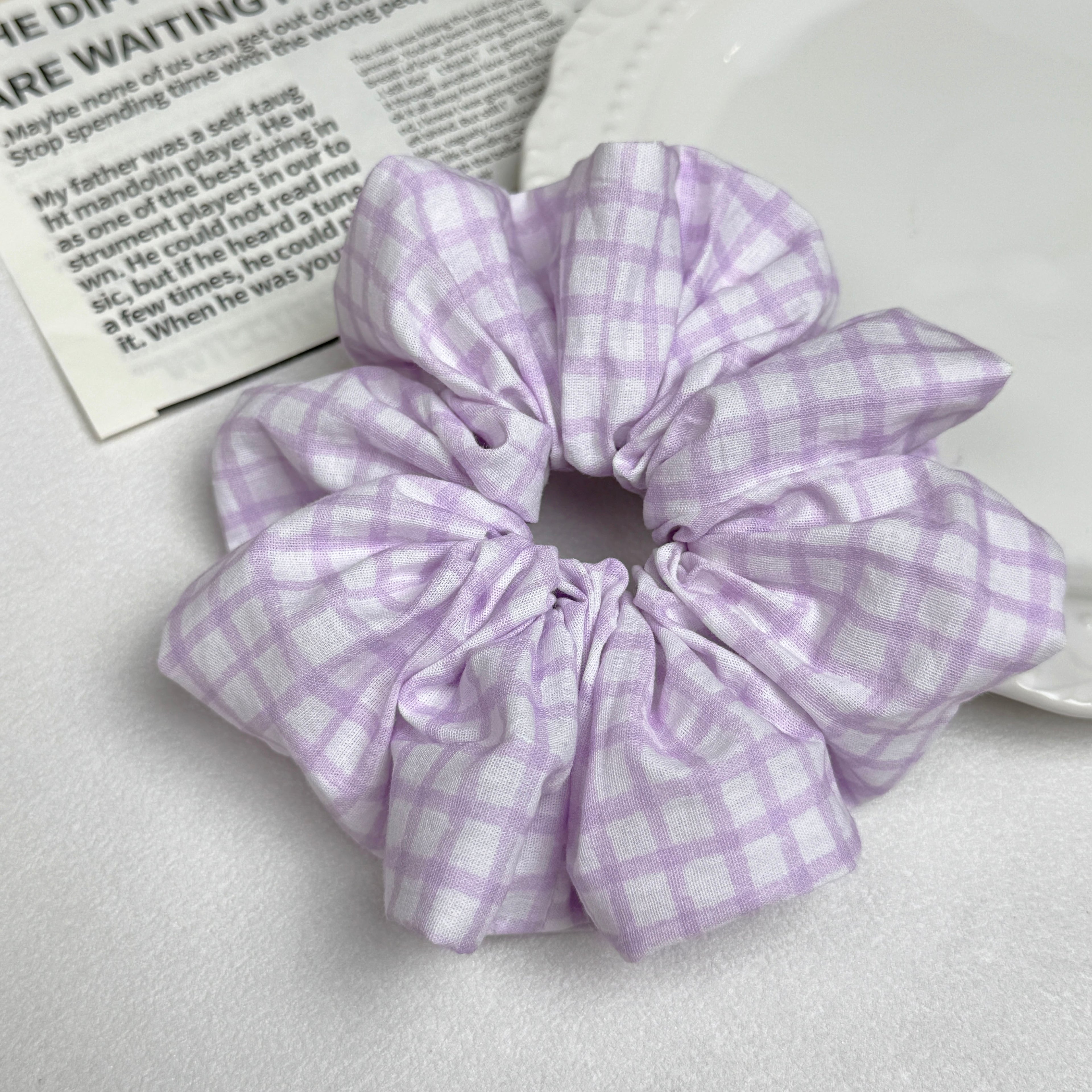 Lavender windowpane plaid scrunchie alongside a white ceramic plate with beaded scalloped rim and an open magazine page with printed text, flat-lay composition.