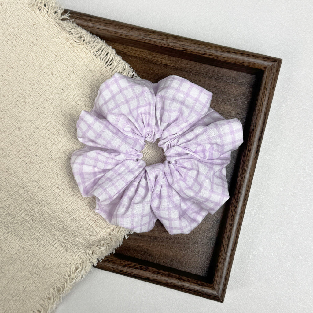 Lavender windowpane plaid scrunchie flat-laid across a dark brown wooden tray and a frayed cream woven fabric with tasseled edges, grey-white textured surface.