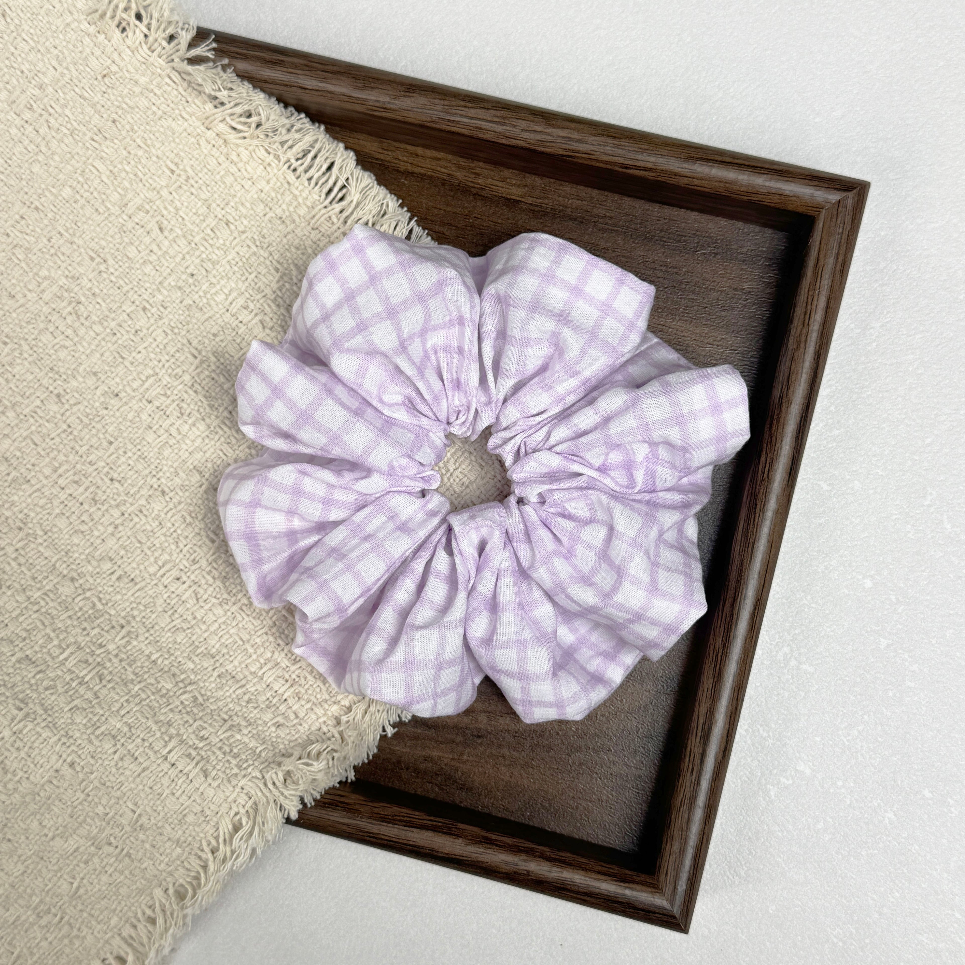 Lavender windowpane plaid scrunchie flat-laid across a dark brown wooden tray and a frayed cream woven fabric with tasseled edges, grey-white textured surface.