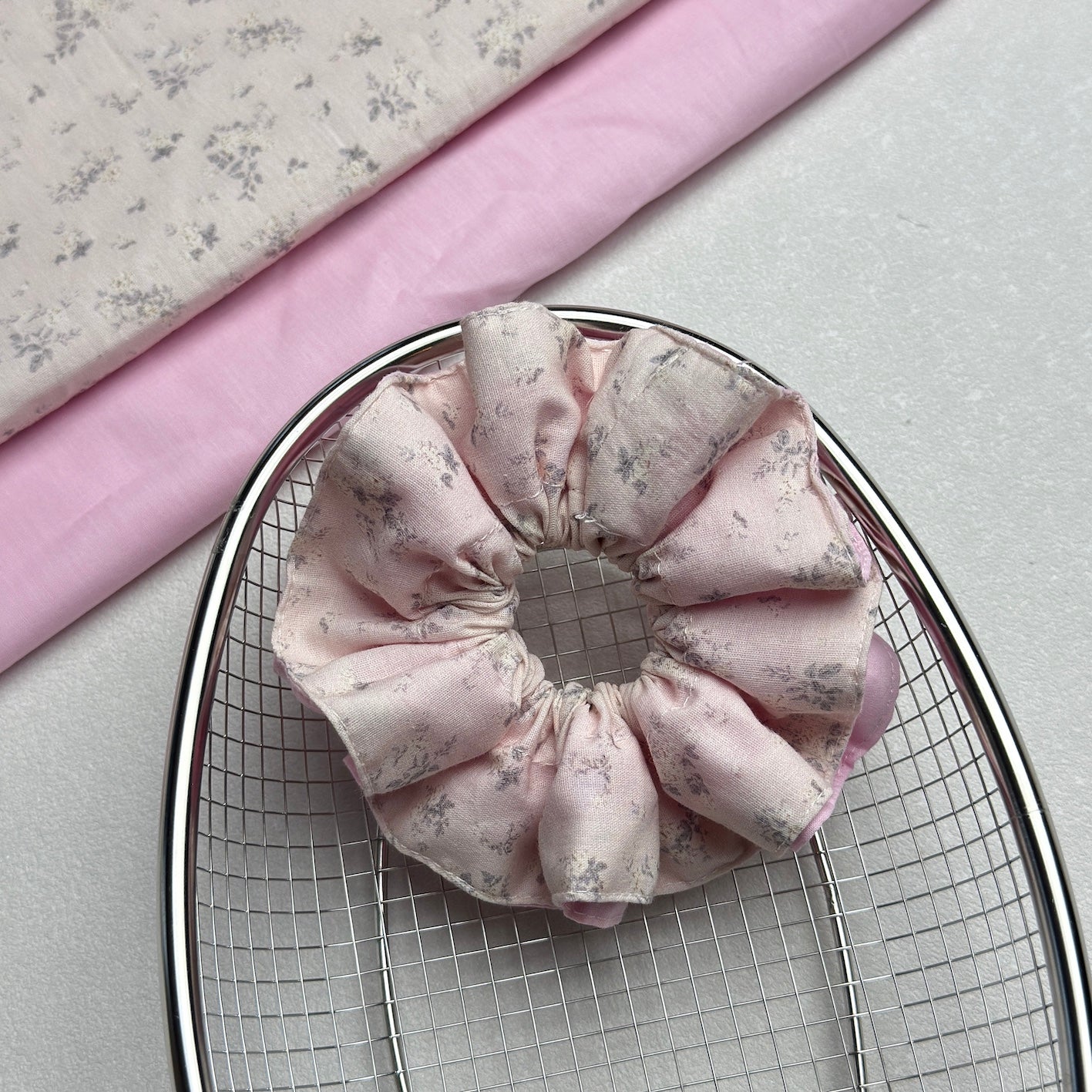 Dusty pink layered floral scrunchie resting in a polished silver wire mesh basket, with matching floral and solid pink folded fabrics in the top left corner, grey-white pebbled surface.