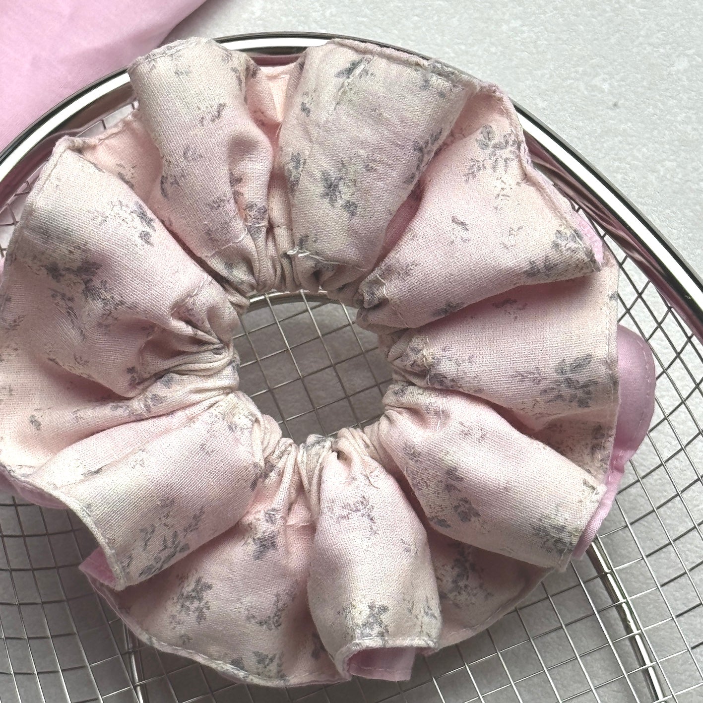 Dusty pink layered floral scrunchie resting on a circular silver wire cooling rack, with a piece of solid pink fabric draped in the top left corner, grey-white textured surface.