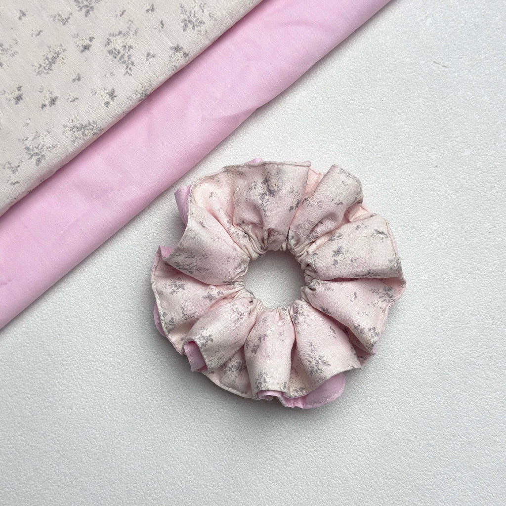 Dusty pink layered floral scrunchie displayed alongside two coordinating fabric pieces — the floral outer fabric and the solid pink underlayer — laid diagonally in the top left corner, white pebbled surface.