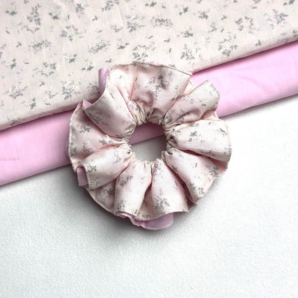 Dusty pink layered floral scrunchie with two neatly folded fabrics — the grey-blue floral print and the solid pink lining — running diagonally behind it, white textured surface.