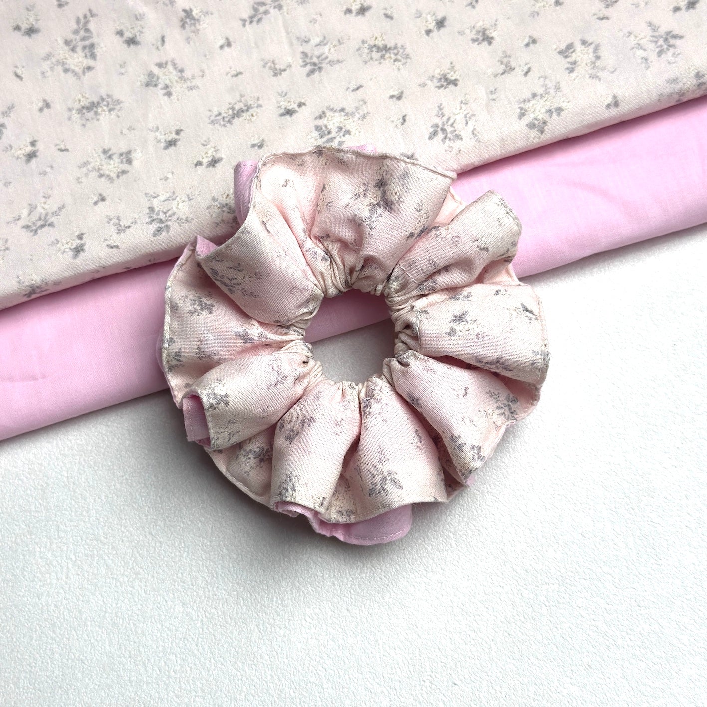 Dusty pink layered floral scrunchie with two neatly folded fabrics — the grey-blue floral print and the solid pink lining — running diagonally behind it, white textured surface.