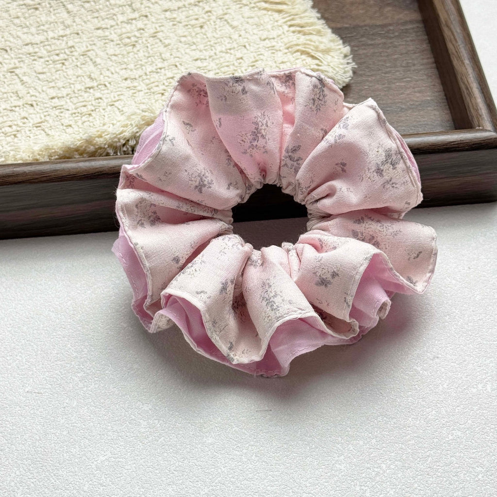 Dusty pink layered floral scrunchie on a grey-white surface, with a dark polished wooden tray and fringed cream waffle-weave cloth in the background, flat-lay composition.