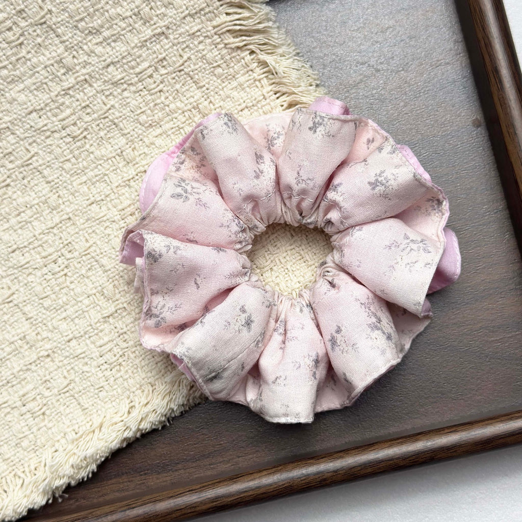 Dusty pink layered floral scrunchie resting on a fringed cream waffle-weave cloth inside a dark brown wooden tray, solid pink underlayer visible at the edges, flat-lay composition.
