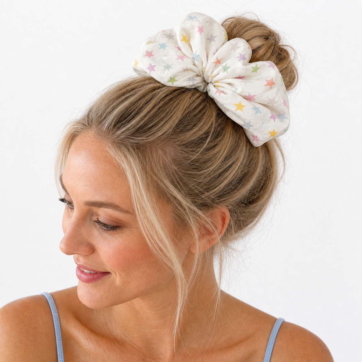 Blonde woman wearing a white pastel star print scrunchie in a high messy bun, side profile, light blue tank top, clean white background.