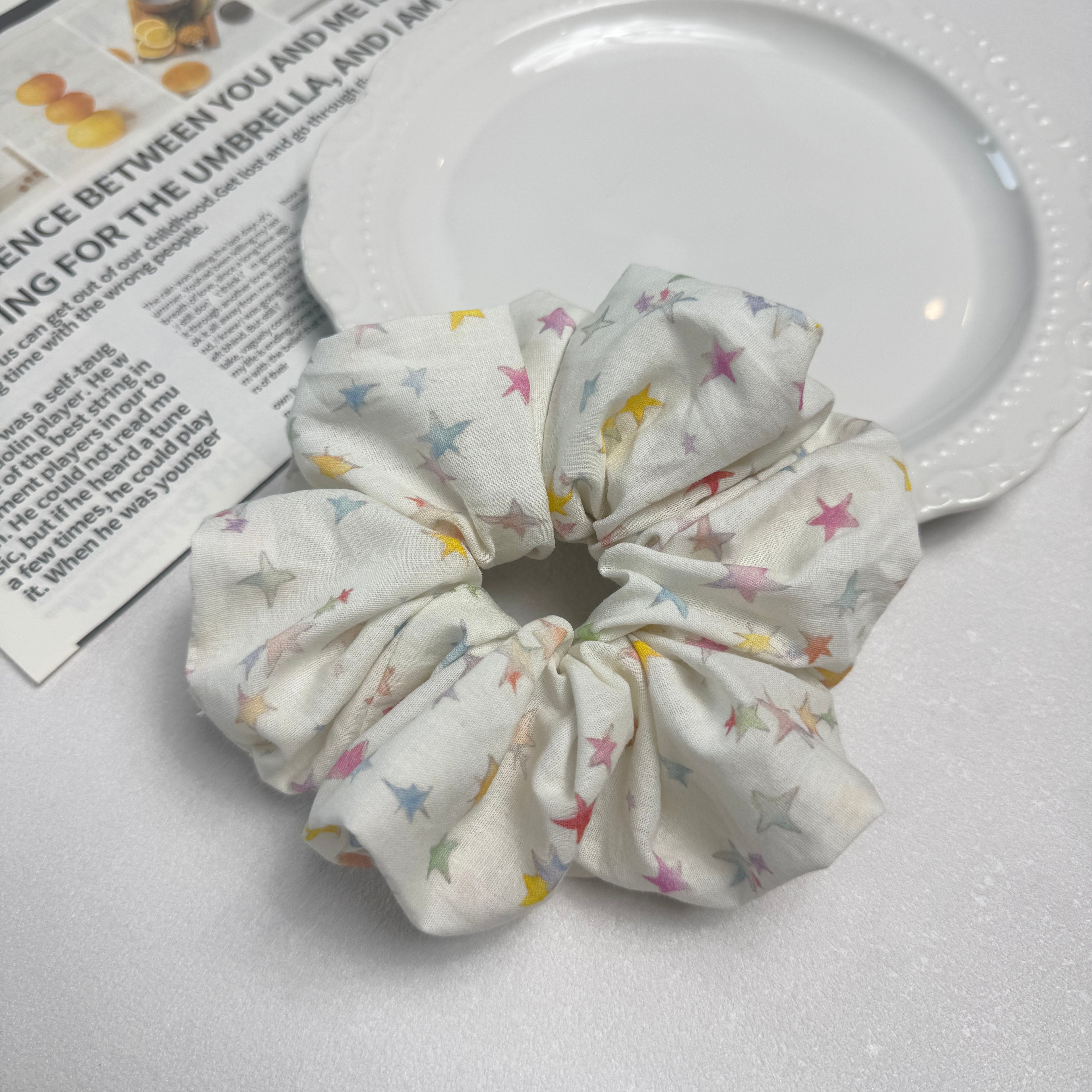 White pastel star print scrunchie alongside a white ceramic plate with beaded rim and a magazine page featuring citrus fruit photography, top-down flat lay, textured white surface.