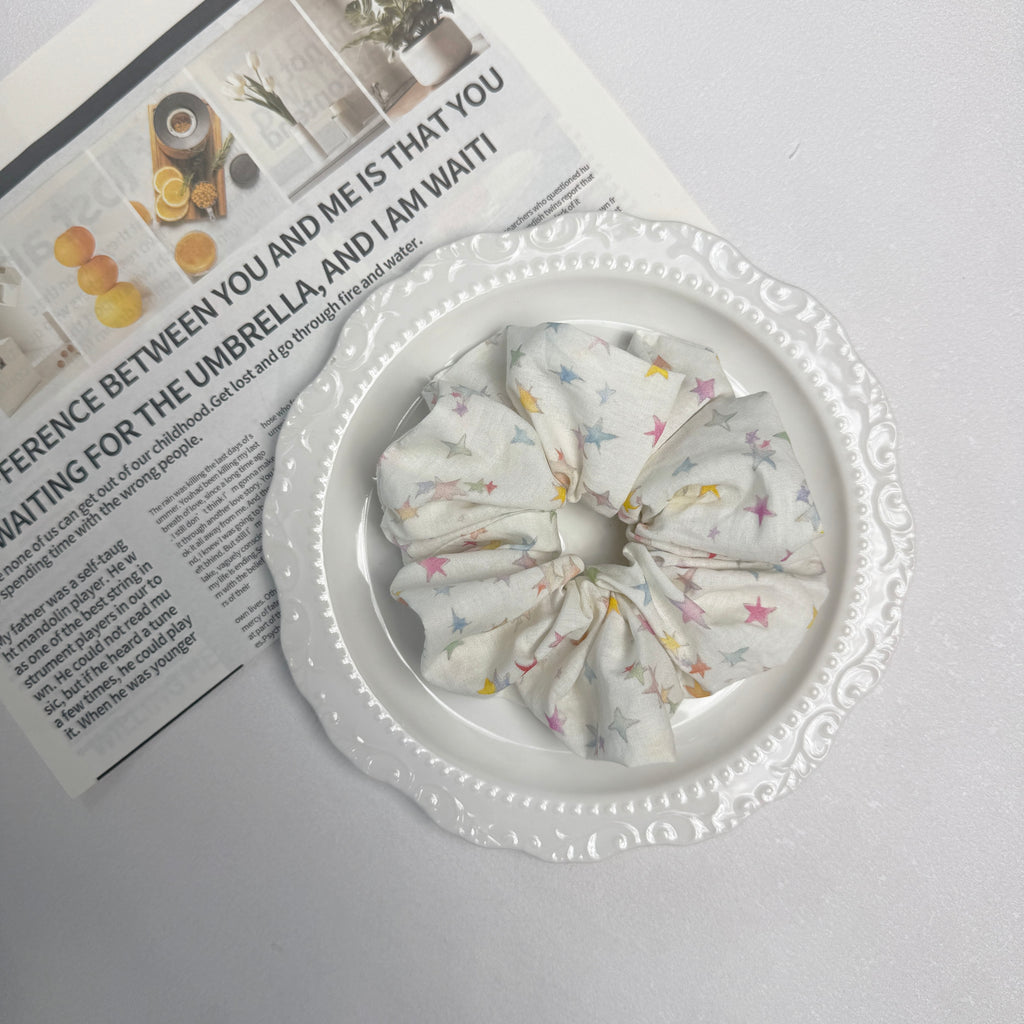 White pastel star print scrunchie placed on a white ceramic plate with embossed scalloped rim, resting on an open magazine page with citrus and home decor photography, grey surface.