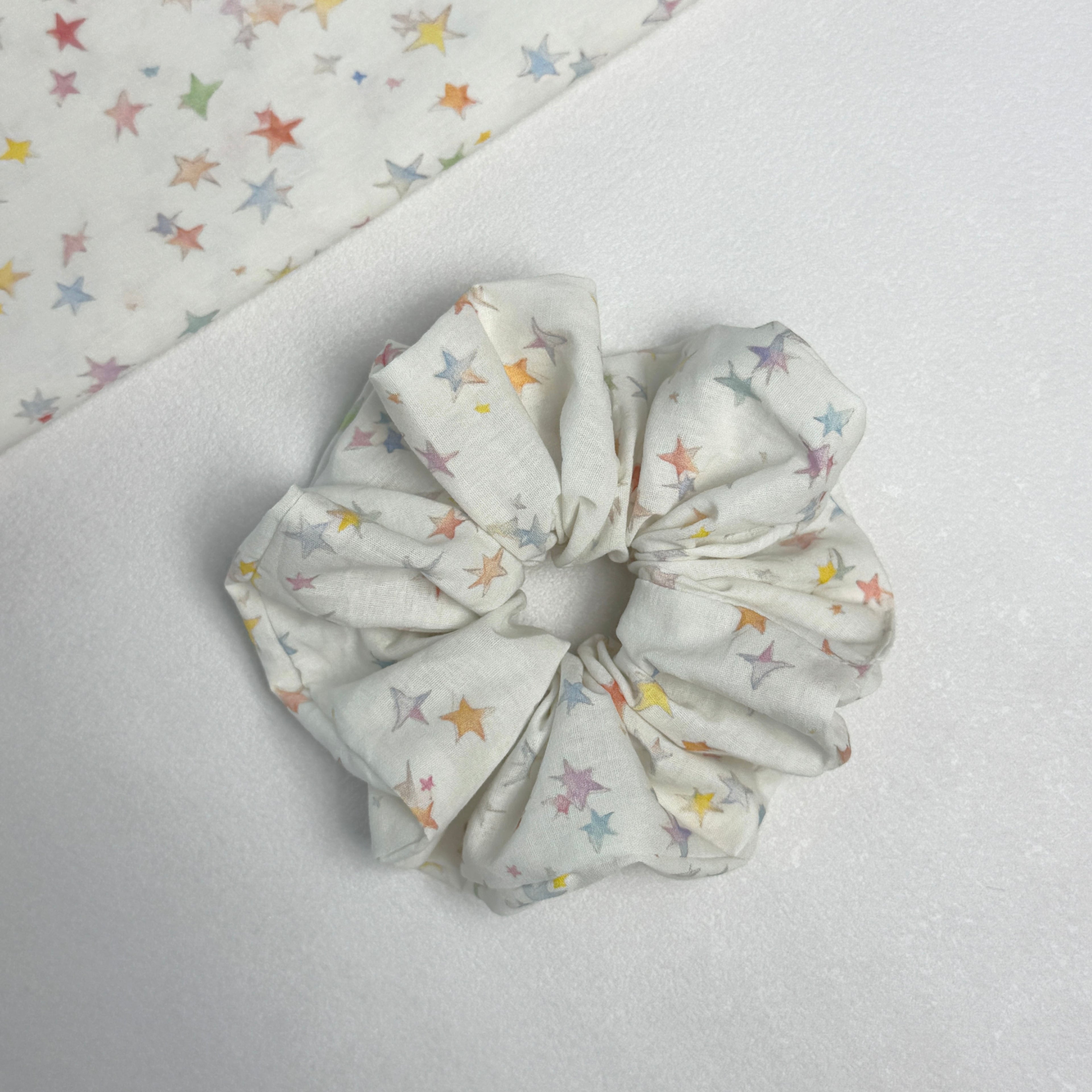 White pastel star print scrunchie displayed alongside a matching flat fabric swatch showing the full multicolor star pattern, white textured surface.
