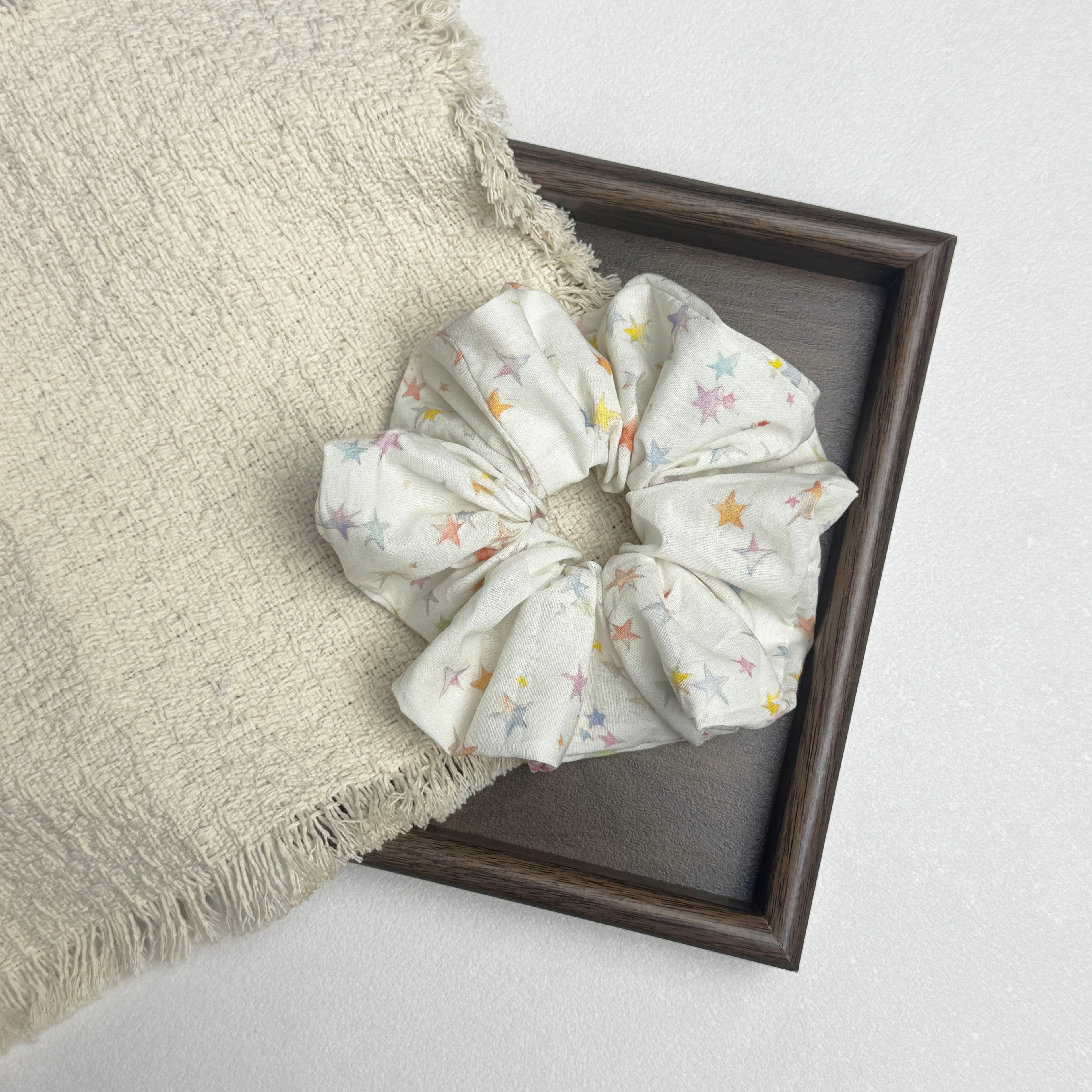 White pastel star print scrunchie flat-laid across a dark brown wooden picture frame and a frayed cream woven fabric, textured grey-white surface.
