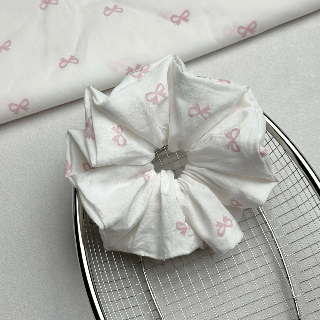 White pink bow print scrunchie resting on a polished silver wire mesh basket, with a matching folded fabric piece in the top left corner, grey-white pebbled surface.