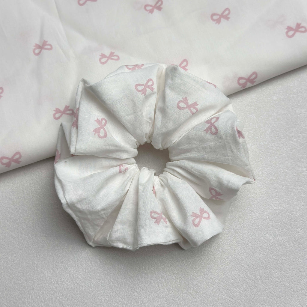 White pink bow print scrunchie with a larger piece of the same fabric draped in the top left corner, off-white grainy surface, flat-lay composition.