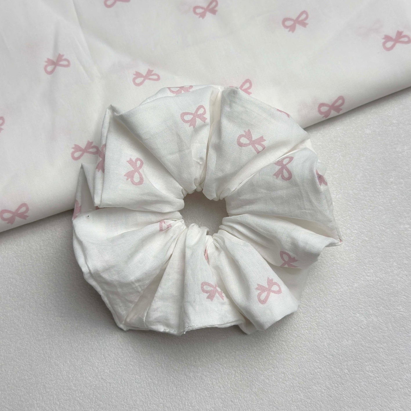 White pink bow print scrunchie with a larger piece of the same fabric draped in the top left corner, off-white grainy surface, flat-lay composition.