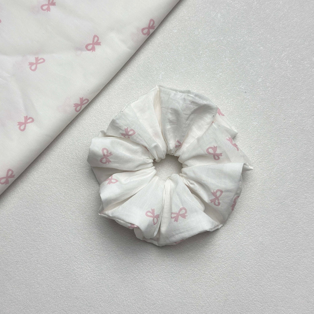White pink bow print scrunchie displayed alongside a matching folded fabric swatch showing the repeating bow pattern, white pebbled texture surface.