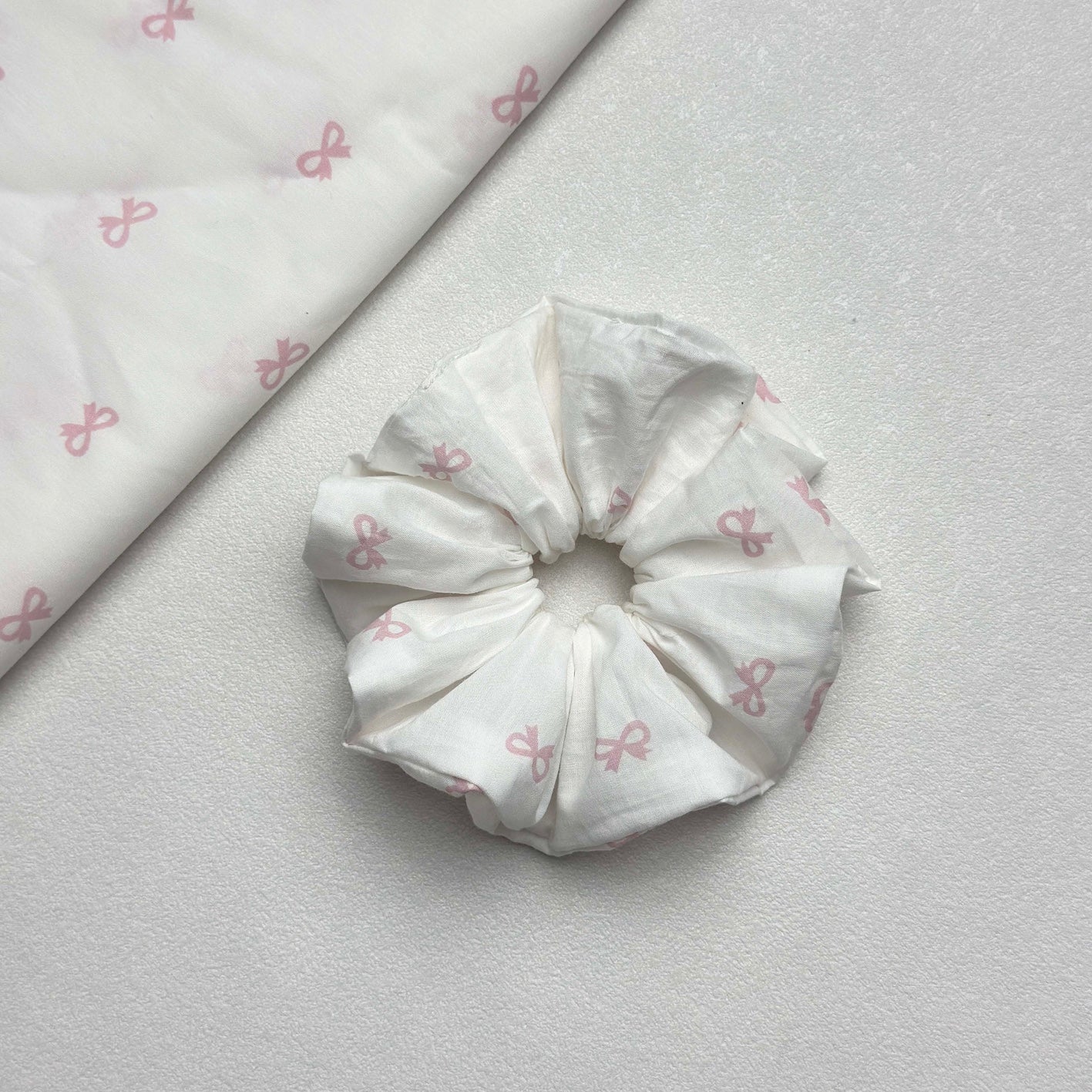 White pink bow print scrunchie displayed alongside a matching folded fabric swatch showing the repeating bow pattern, white pebbled texture surface.
