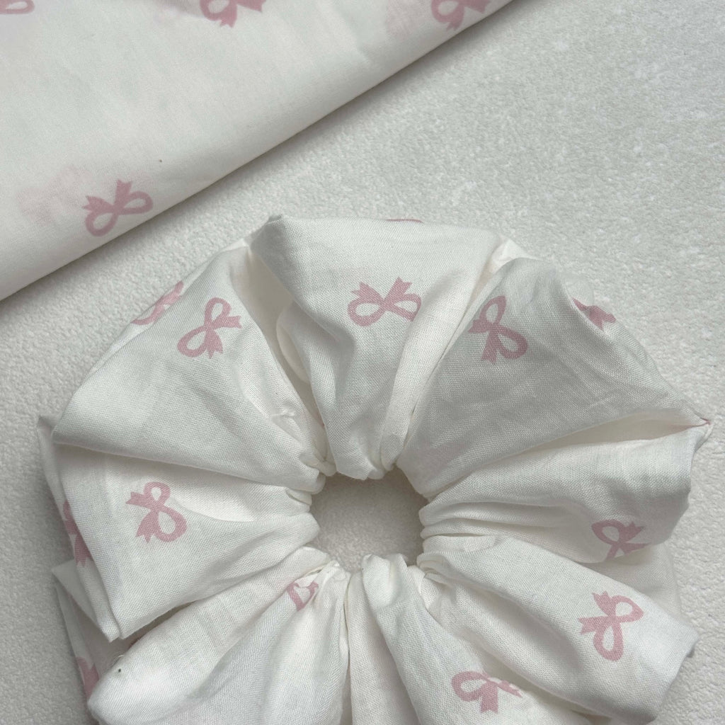White pink bow print scrunchie with a matching folded fabric piece entering diagonally from the top left corner, white pebbled texture surface, flat-lay composition.