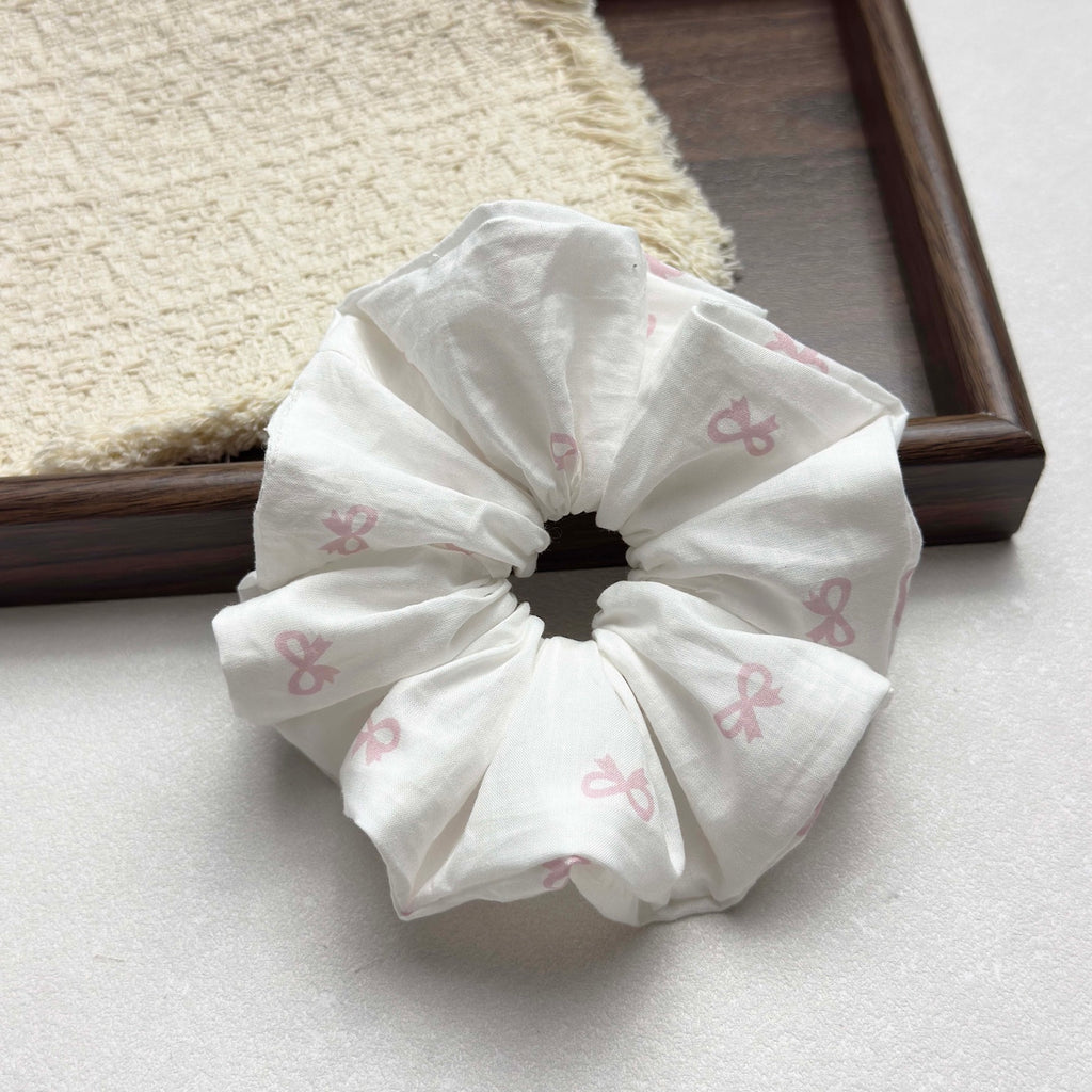 White pink bow print scrunchie on a grey-white surface with a dark brown wooden tray running diagonally and a fringed cream waffle-weave cloth in the top left corner, flat-lay composition.