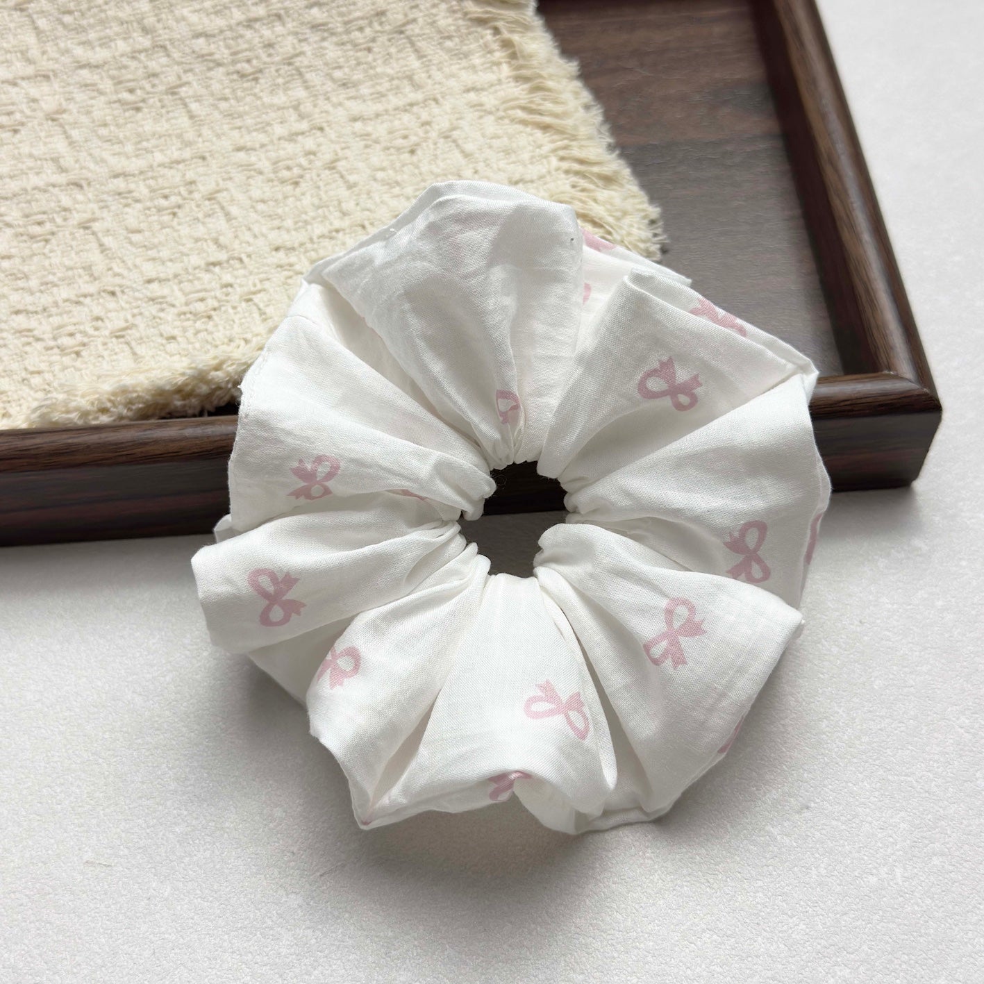 White pink bow print scrunchie on a grey-white surface with a dark brown wooden tray running diagonally and a fringed cream waffle-weave cloth in the top left corner, flat-lay composition.