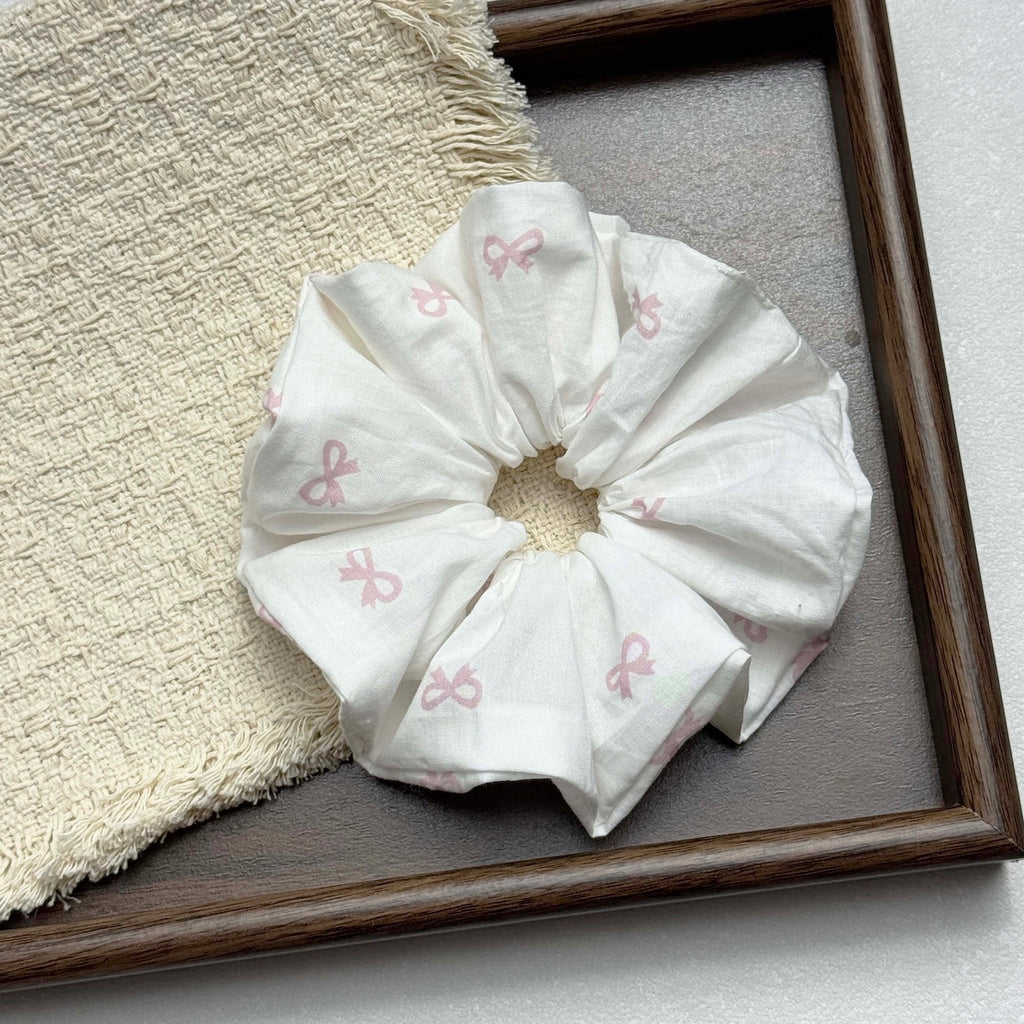 White pink bow print scrunchie resting on a fringed cream waffle-weave cloth inside a dark brown wooden tray with pebbled inner surface, grey-white surface.