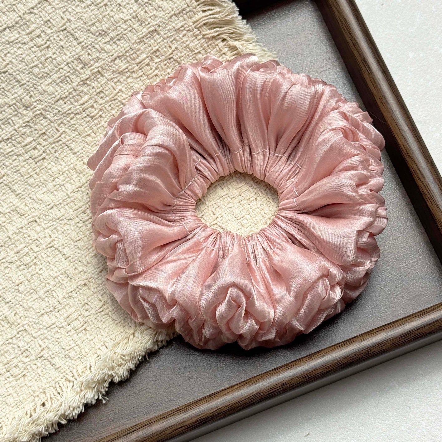 Pink organza cloud scrunchie resting on a fringed cream woven textile inside a dark grey textured tray, grey-white surface, flat-lay composition.