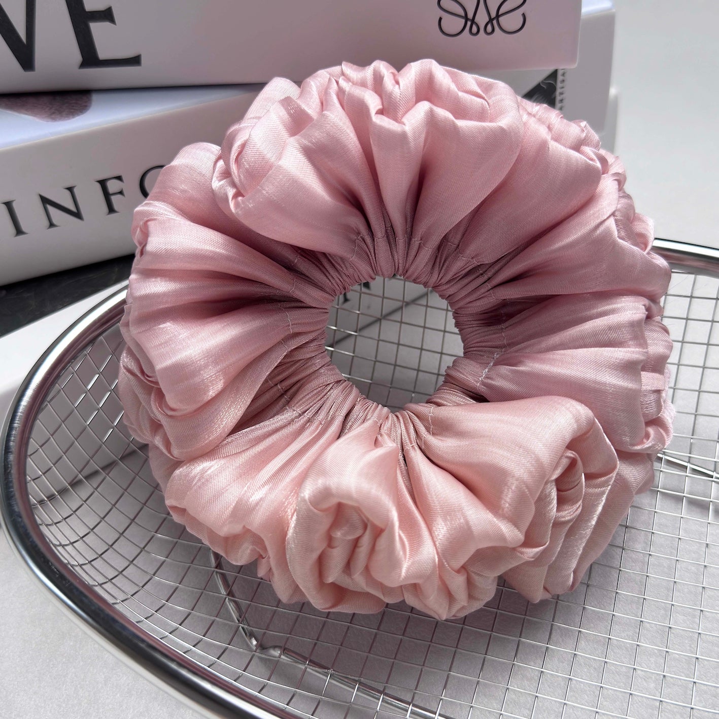 Pink organza cloud scrunchie resting in a polished silver wire mesh basket, placed on stacked white art and lifestyle books, light grey surface.