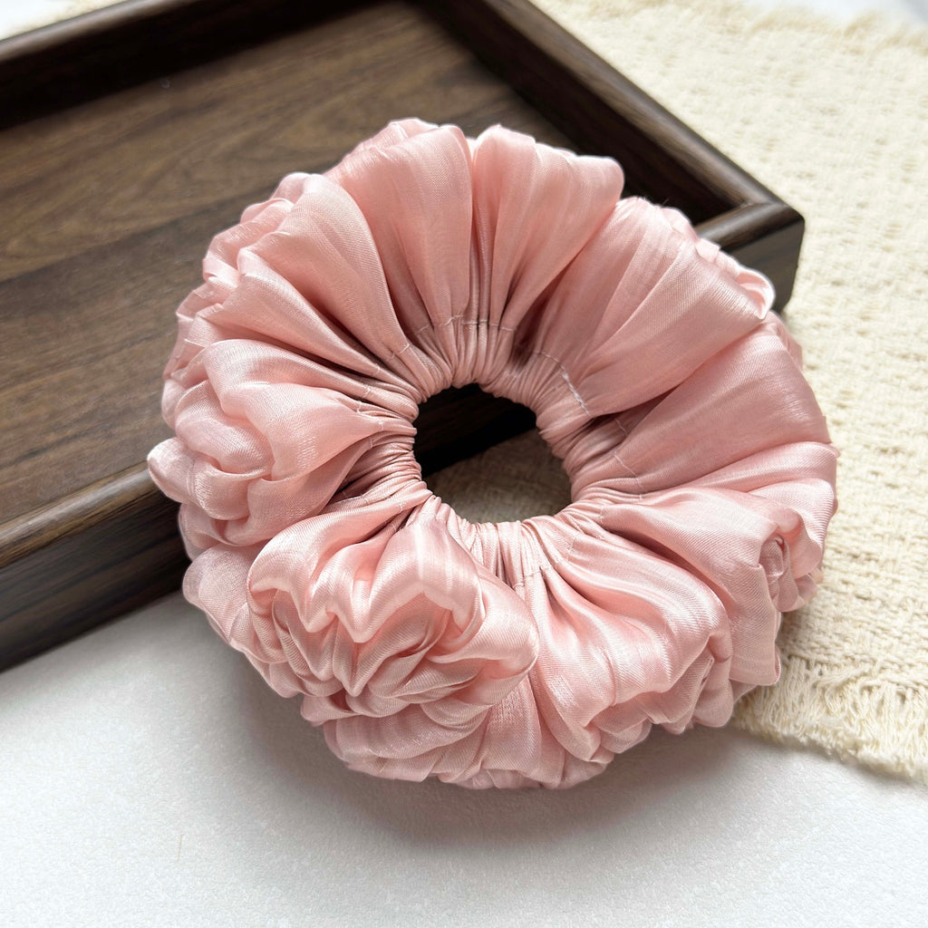Pink organza cloud scrunchie on a white textured surface, with a dark brown wooden tray corner in the background and a fringed cream woven fabric to the right, flat-lay composition.