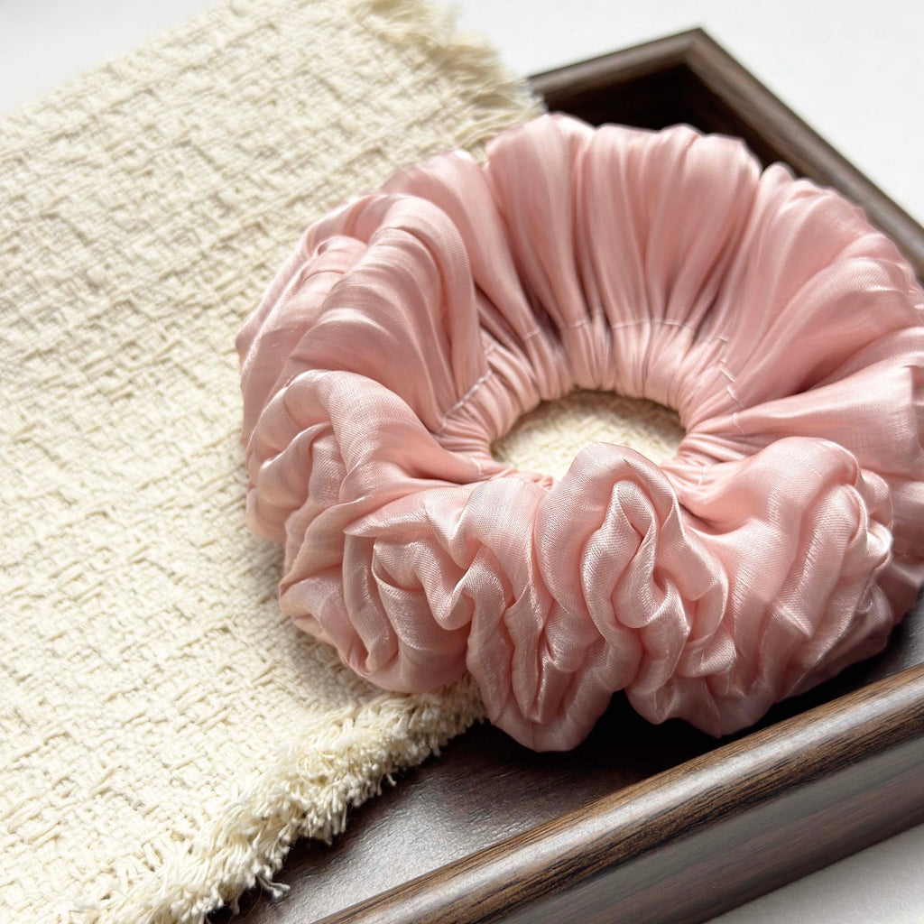 Pink organza cloud scrunchie resting on a fringed cream woven textile inside a dark brown wooden tray with visible grain, flat-lay composition.