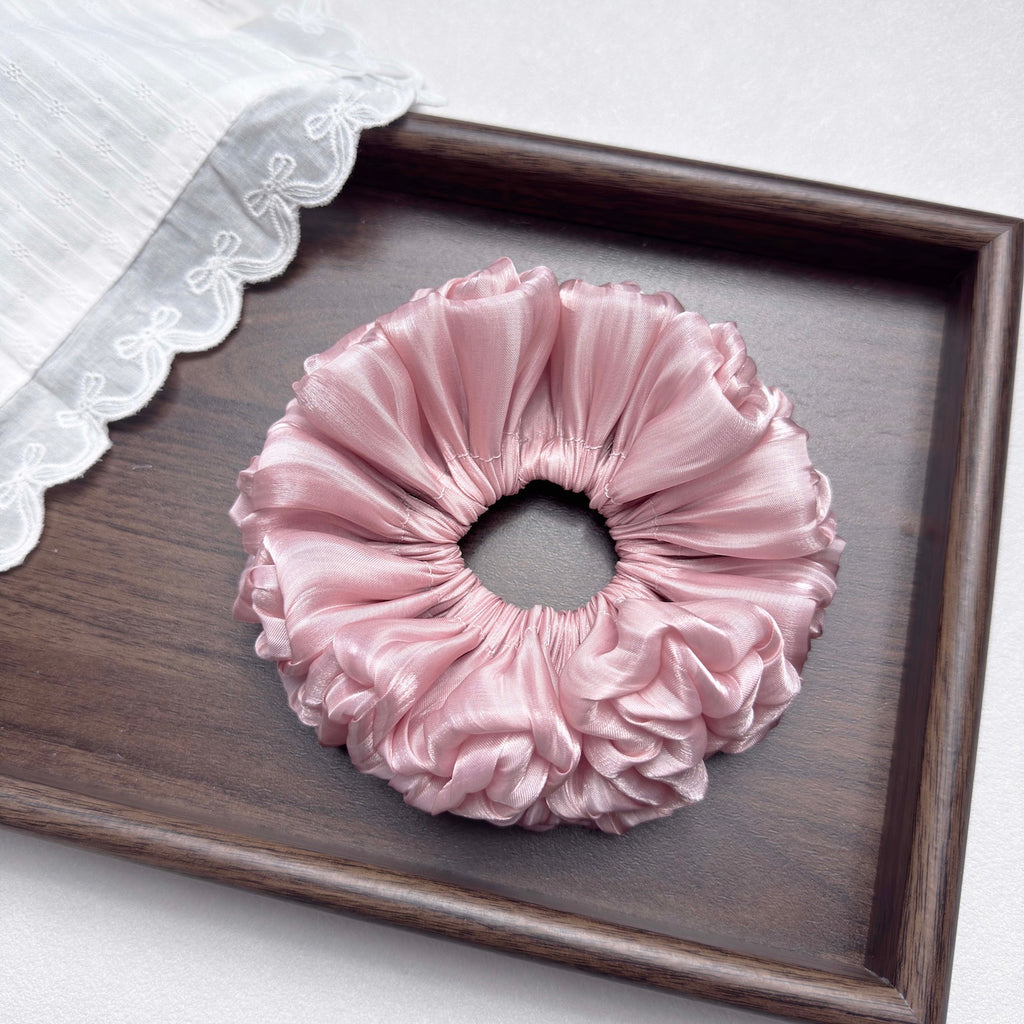 Pink organza cloud scrunchie centered in a dark brown wooden tray, a piece of white striped fabric with delicate eyelet lace trim draped over the top left corner, white surface.