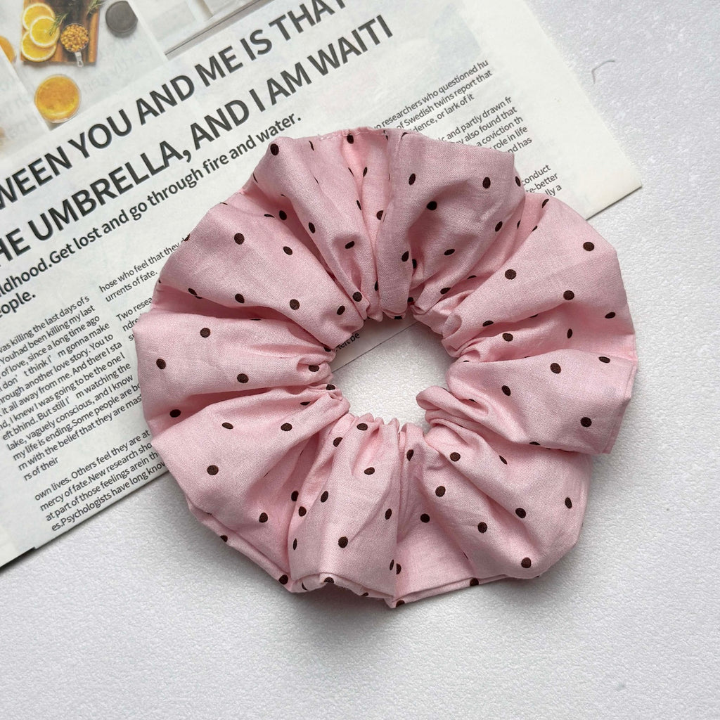 Baby pink polka dot scrunchie resting on a magazine page with printed text and citrus photography, white textured surface, flat-lay composition.