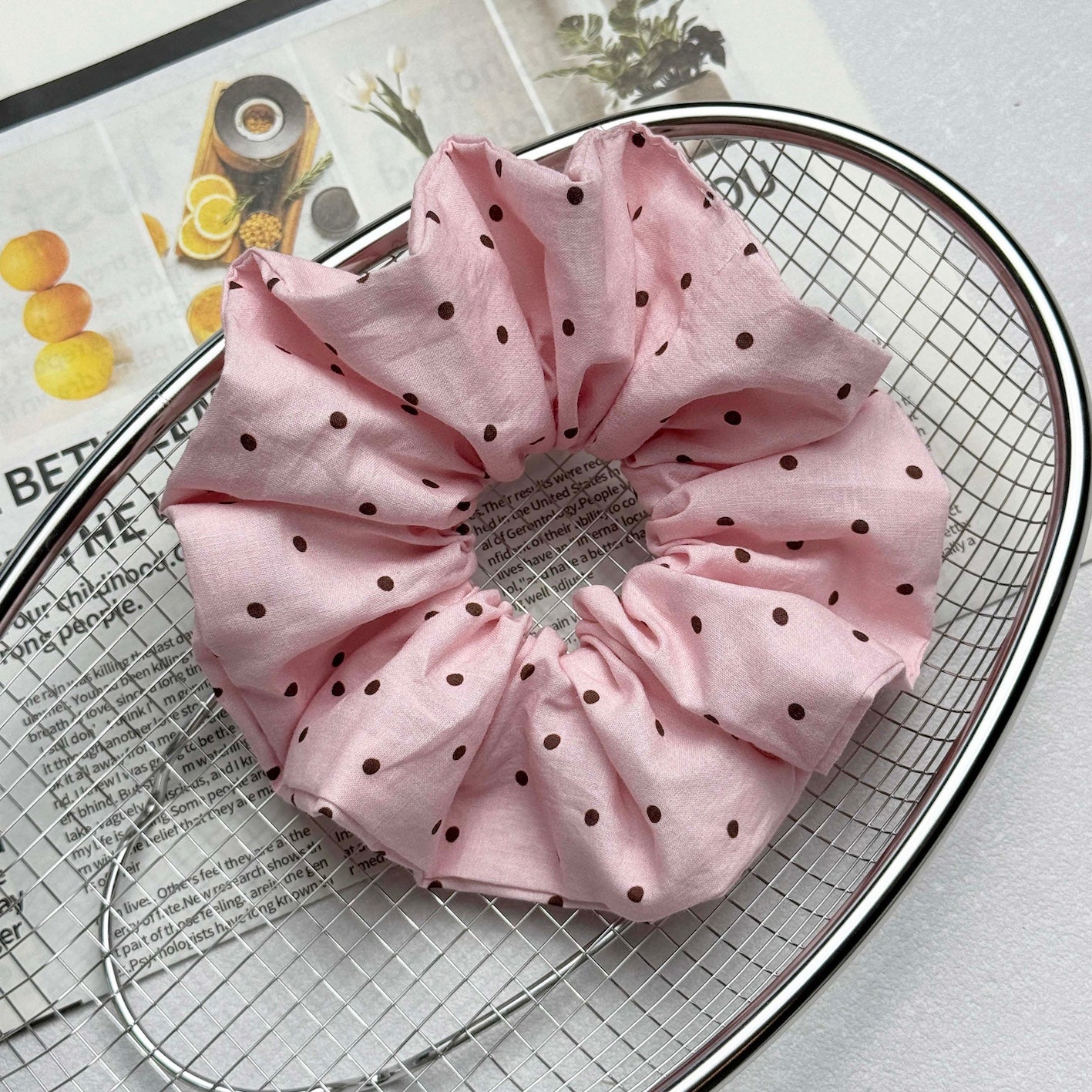 Baby pink polka dot scrunchie resting in a silver oval wire mesh basket, with a magazine page of text and botanical images visible beneath the mesh, white textured surface.