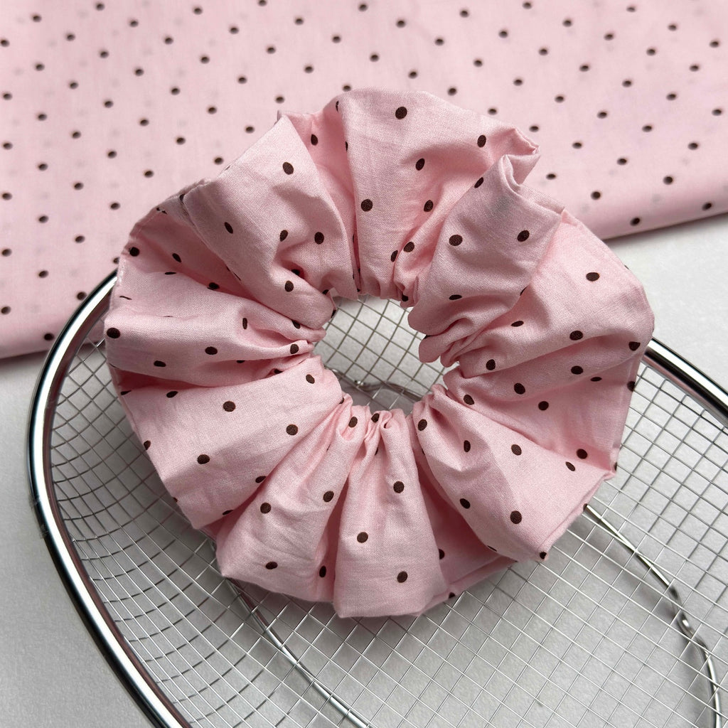 Baby pink polka dot scrunchie resting on a circular chrome wire mesh tray, with a large piece of matching polka dot fabric draped behind it, white textured surface.