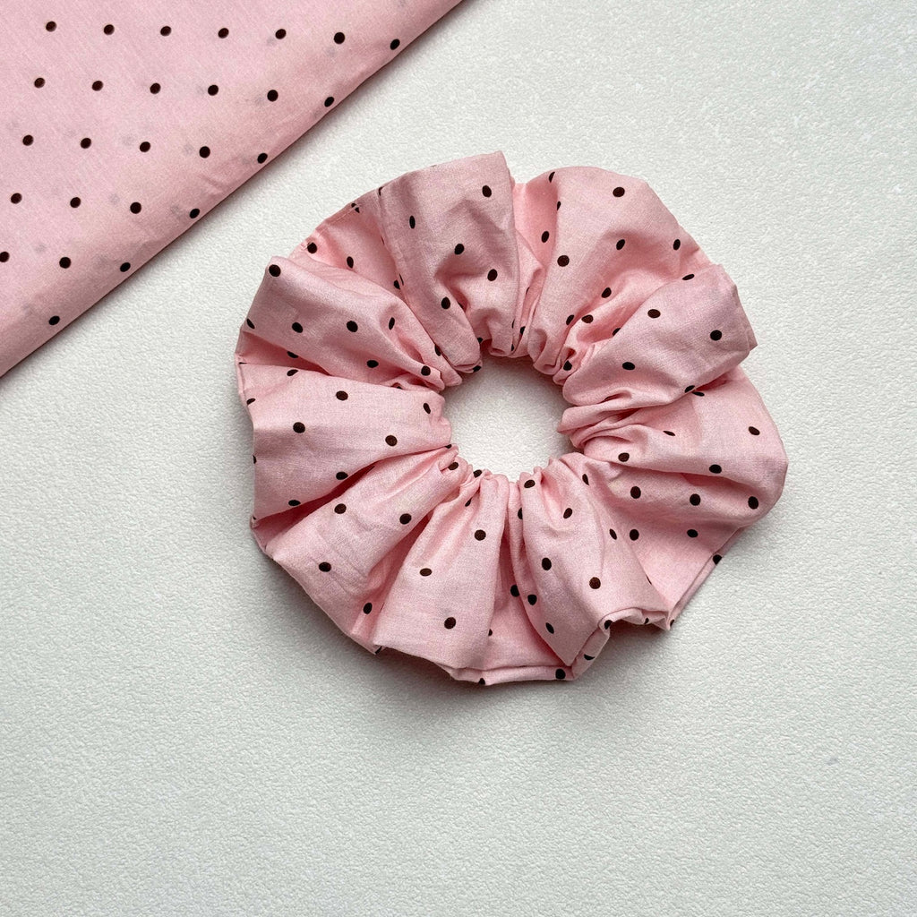 Baby pink polka dot scrunchie displayed alongside a matching fabric swatch entering diagonally from the top left, showing the full dot pattern, white pebbled texture surface.