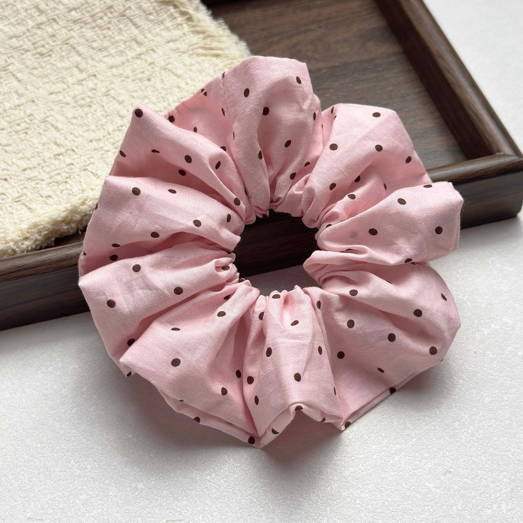 Baby pink polka dot scrunchie on a grey-white pebbled surface, with a dark brown wooden frame running diagonally and a cream waffle-knit cloth in the top left corner.