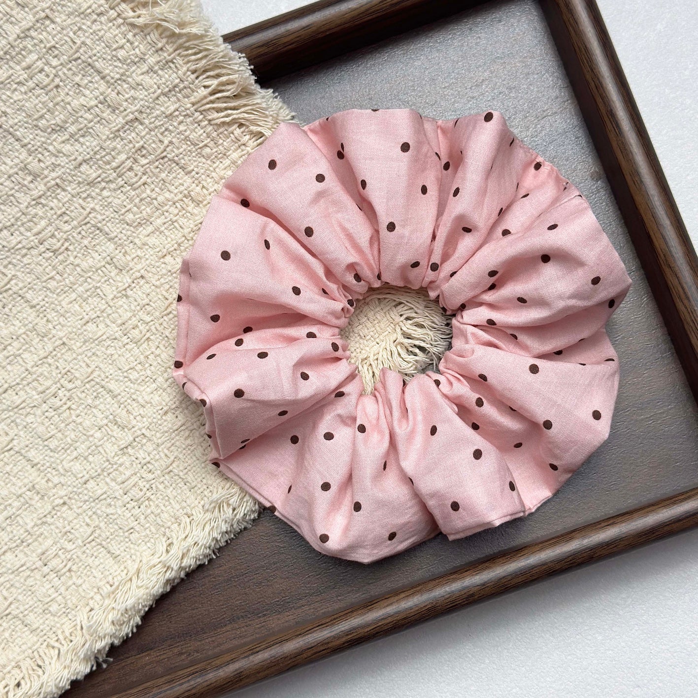 Baby pink polka dot scrunchie resting on a fringed cream woven textile inside a dark brown wooden tray with visible grain, grey-white surface.