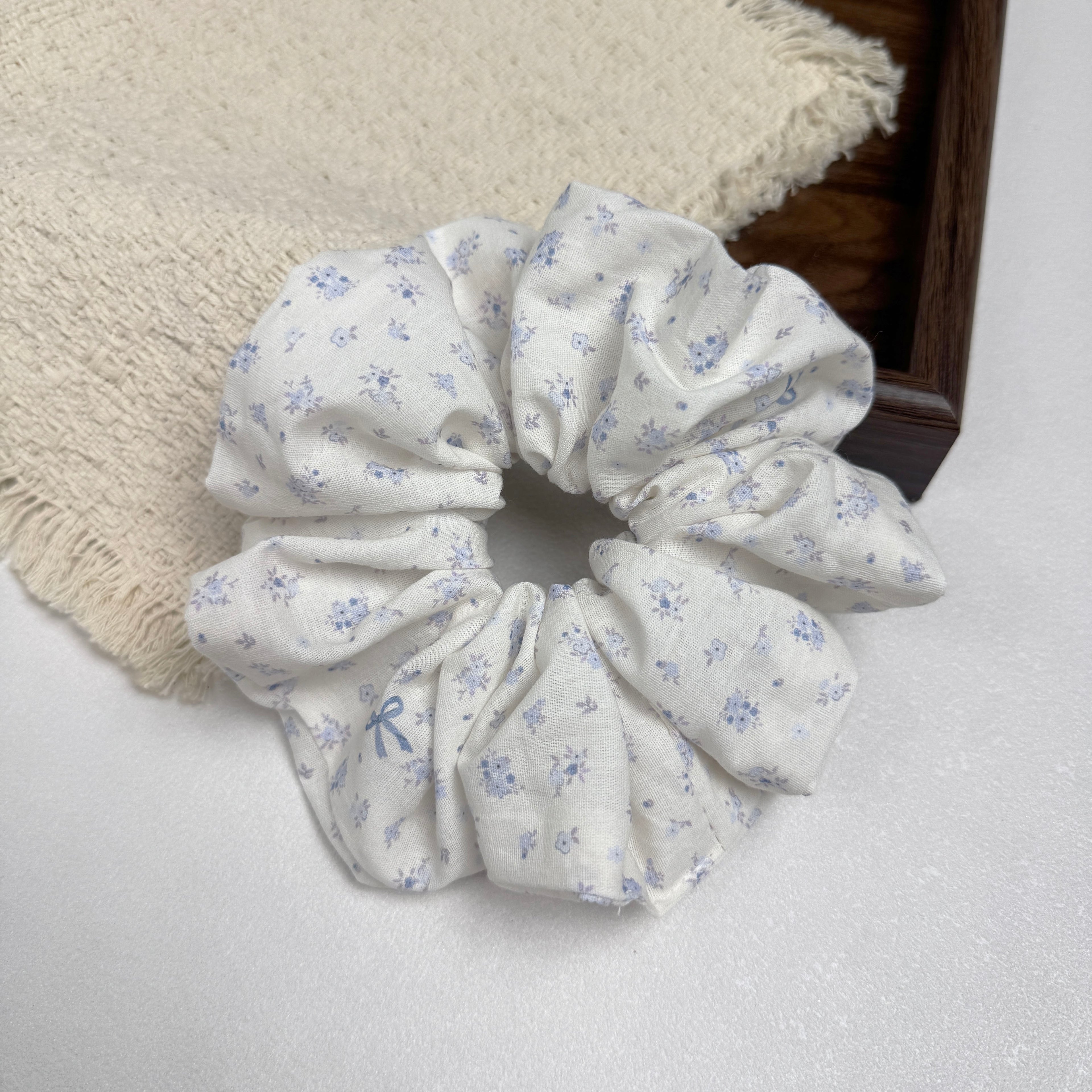 Ivory purple floral bow print scrunchie on a grey-white surface beside a frayed cream woven fabric and the corner of a dark brown wooden tray, flat-lay composition.