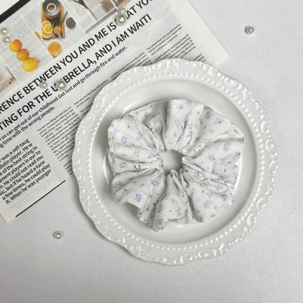 Ivory purple floral bow print scrunchie placed on a white ceramic plate with embossed scalloped rim, resting on a magazine page with citrus photography and text, scattered white faux pearls, grey-white surface.
