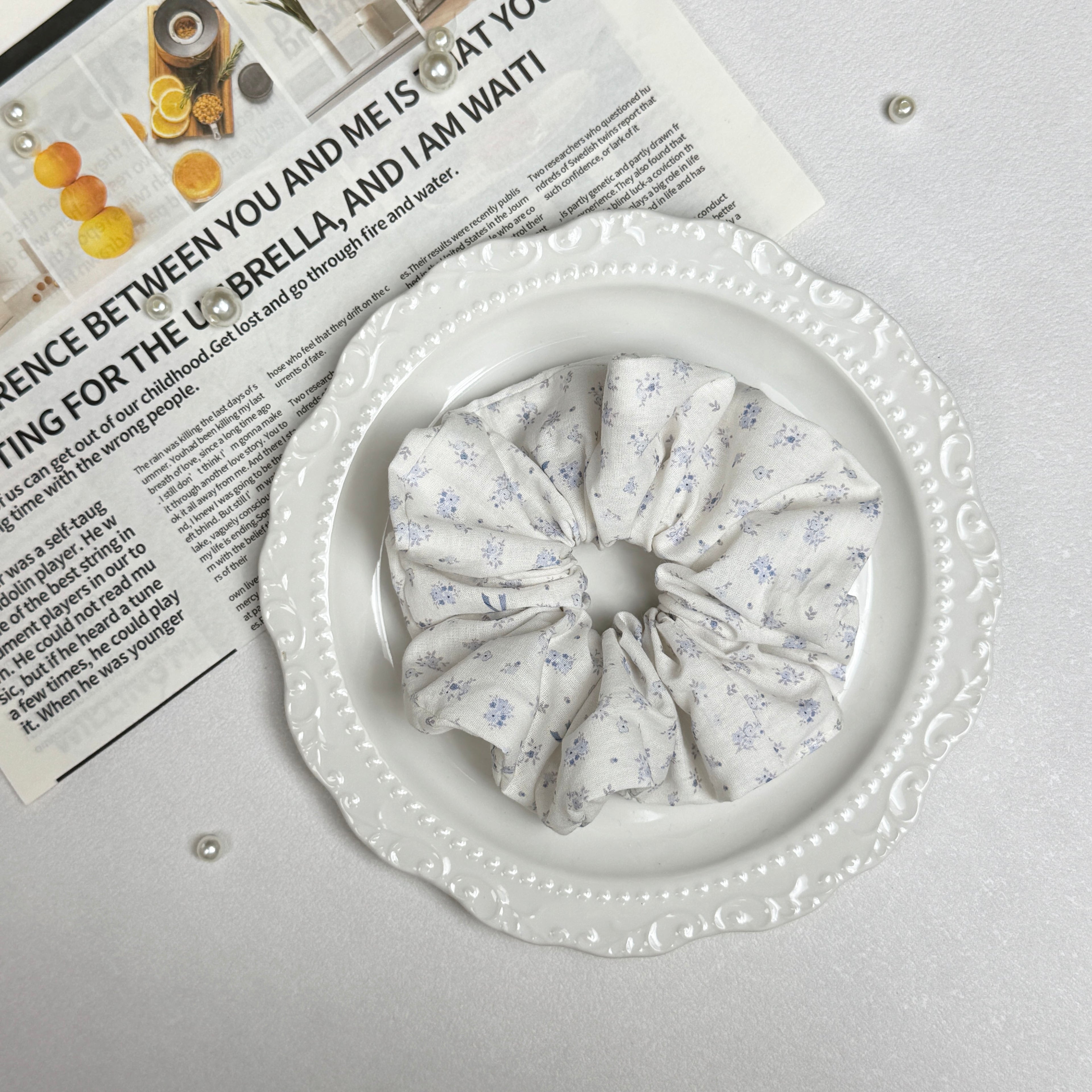 Ivory purple floral bow print scrunchie placed on a white ceramic plate with embossed scalloped rim, resting on a magazine page with citrus photography and text, scattered white faux pearls, grey-white surface.