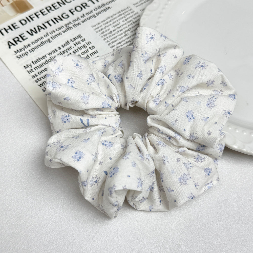 Ivory purple floral bow print scrunchie alongside a white ceramic plate with beaded scalloped rim and an open magazine page with printed text, clean white surface, flat-lay composition.