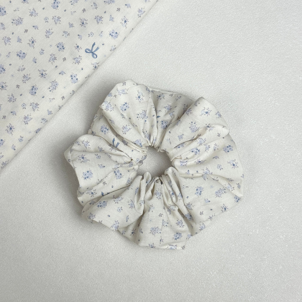 Ivory purple floral bow print scrunchie displayed alongside a matching folded fabric swatch, showing the full floral and bow motif pattern clearly, white pebbled texture surface.