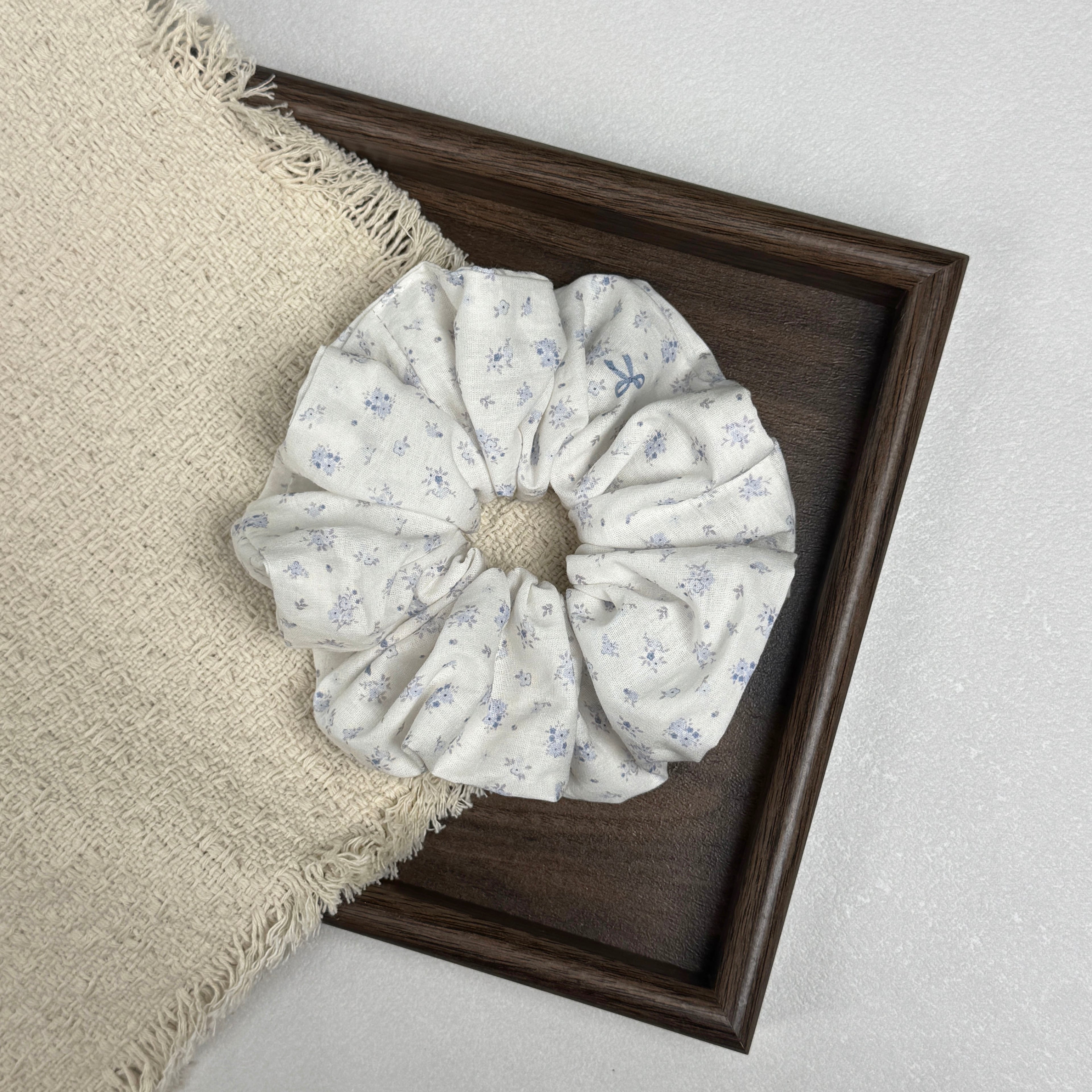Ivory purple floral bow print scrunchie resting partially inside a dark brown wooden tray and partially on a fringed cream woven fabric, grey-white textured surface, flat-lay composition.