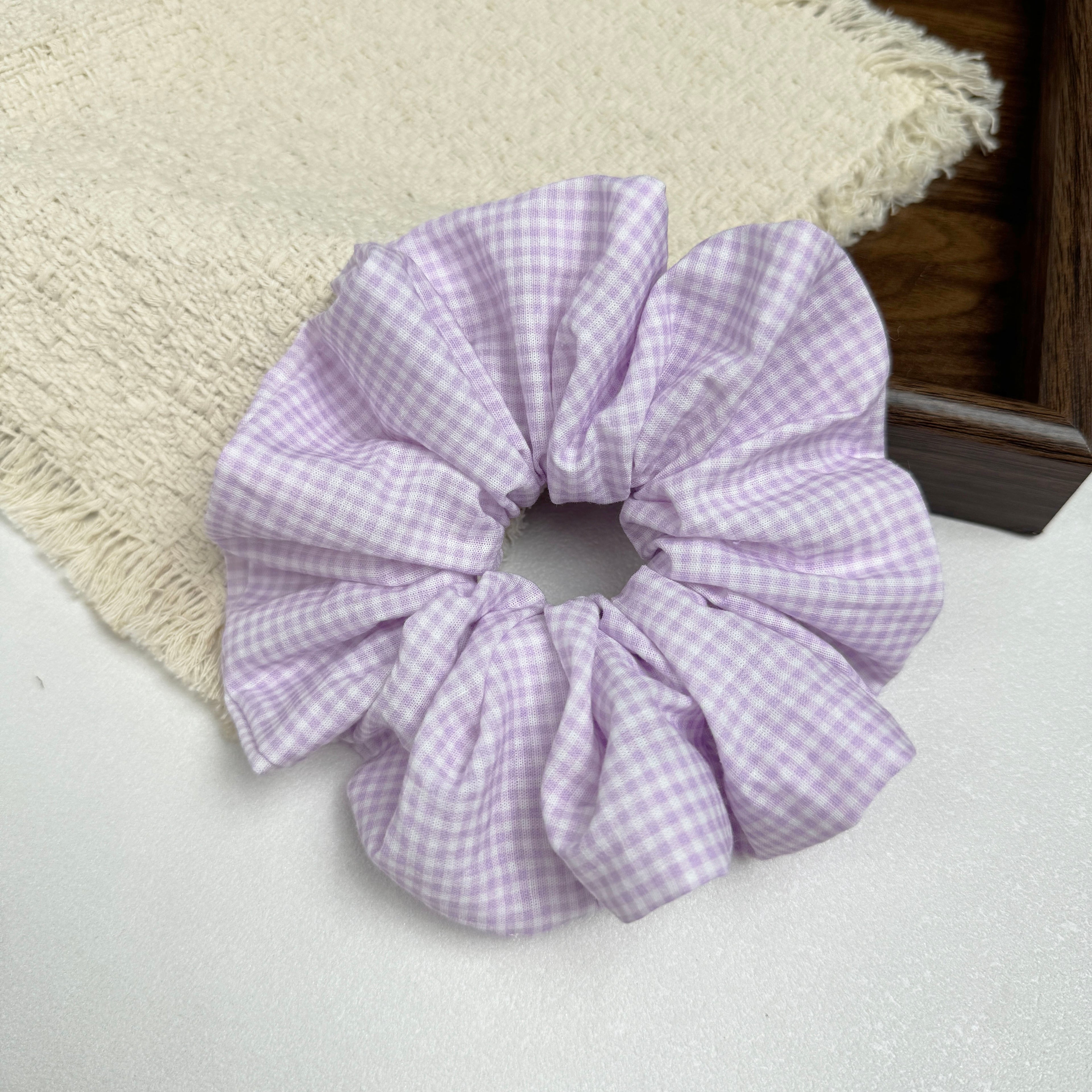 Lavender gingham scrunchie on a grey-white surface beside a frayed cream woven fabric and the corner of a dark brown wooden tray, flat-lay composition.