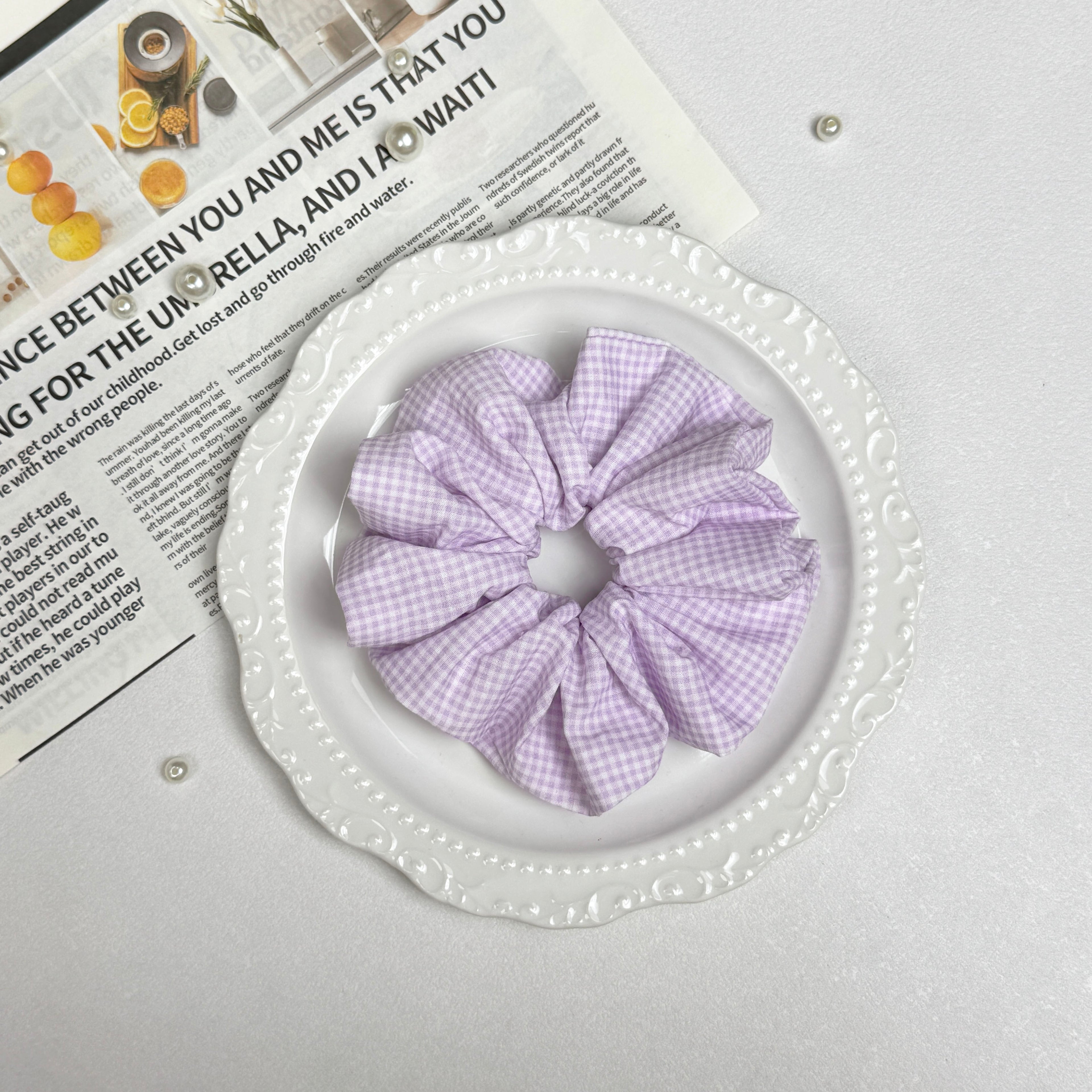 Lavender gingham scrunchie placed on a white ceramic plate with embossed scalloped rim, resting on a magazine page with food photography, scattered white faux pearls on the page, grey-white surface.